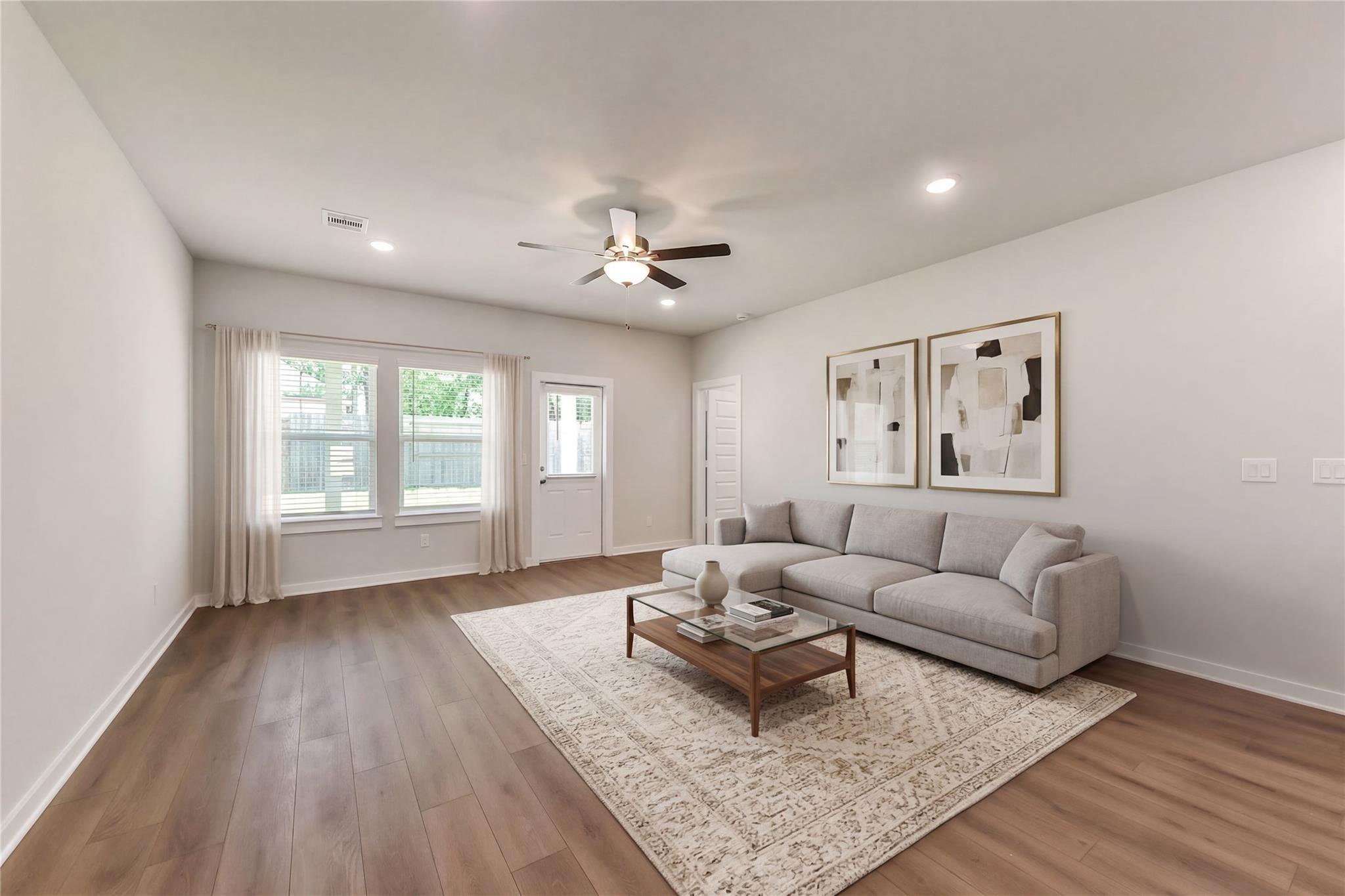 Bright living room featuring gray sectional sofa, coffee table, ceiling fan, and large windows in Davidson Homes The Brazos E, Magnolia Texas