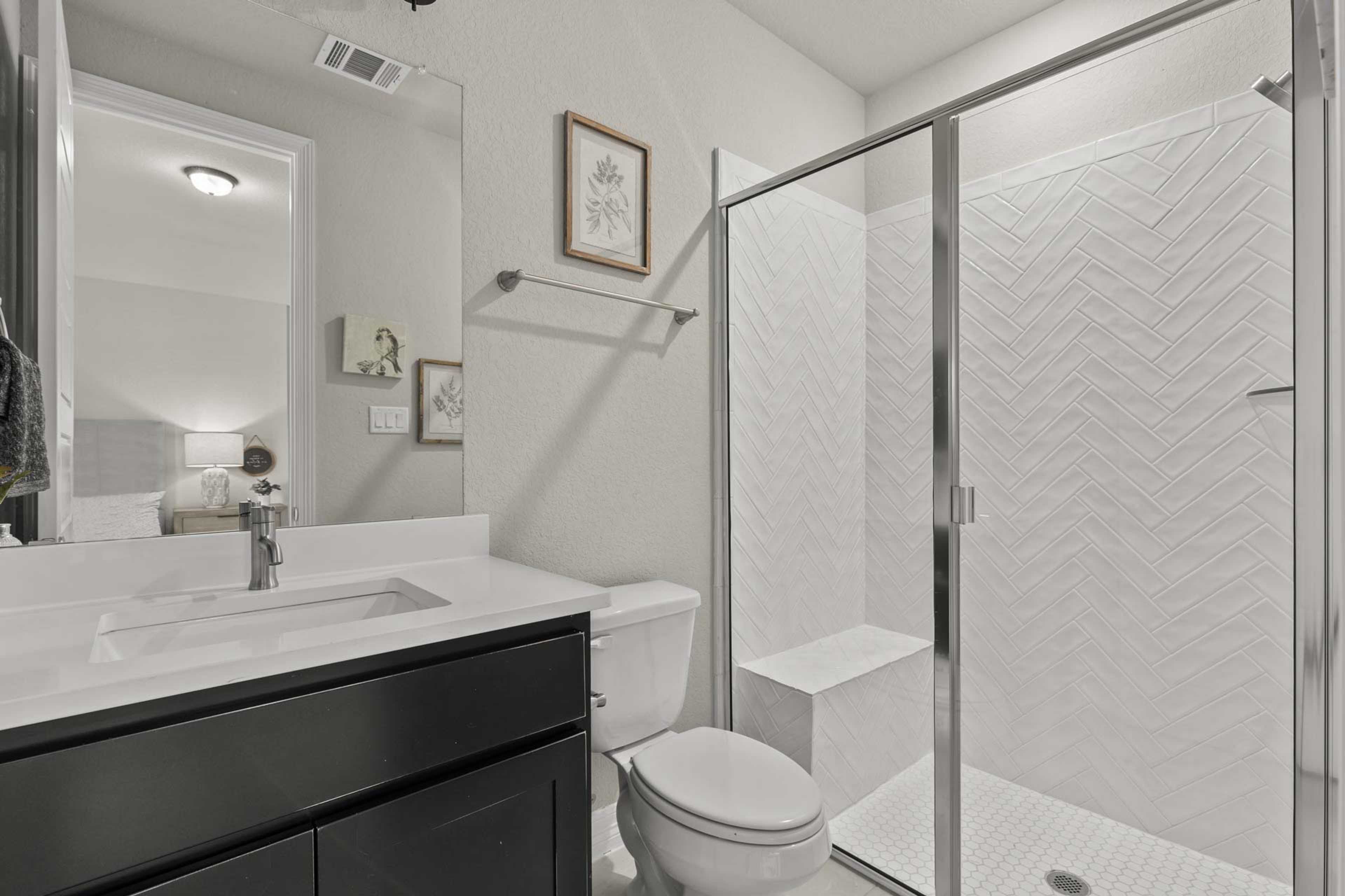Modern bathroom in Potranco Oaks, Castroville TX with white herringbone tile shower, glass enclosure, dark vanity and sleek fixtures by Davidson Homes