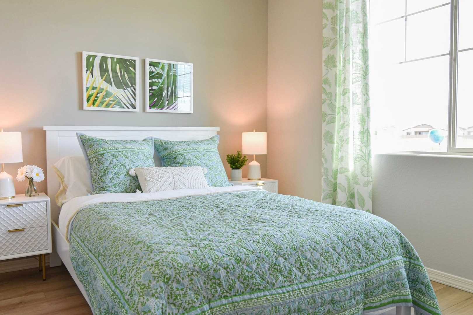 Spacious master bedroom in The Harmony with green quilted bed, palm leaf wall art, sheer curtains, and soft lamp lighting