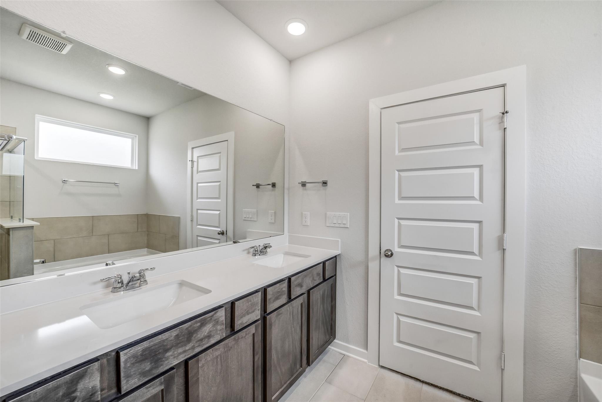 Elegant master bathroom with double vanity, gray cabinets, soaking tub, and frosted window in Davidson Homes The Acadia C, Magnolia Texas