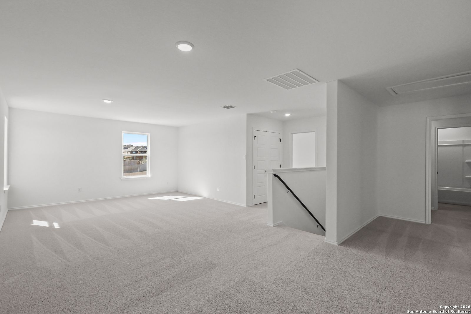 Spacious upstairs loft with white walls, gray carpet, large window, and staircase in Davidson Homes The Douglas E, San Antonio