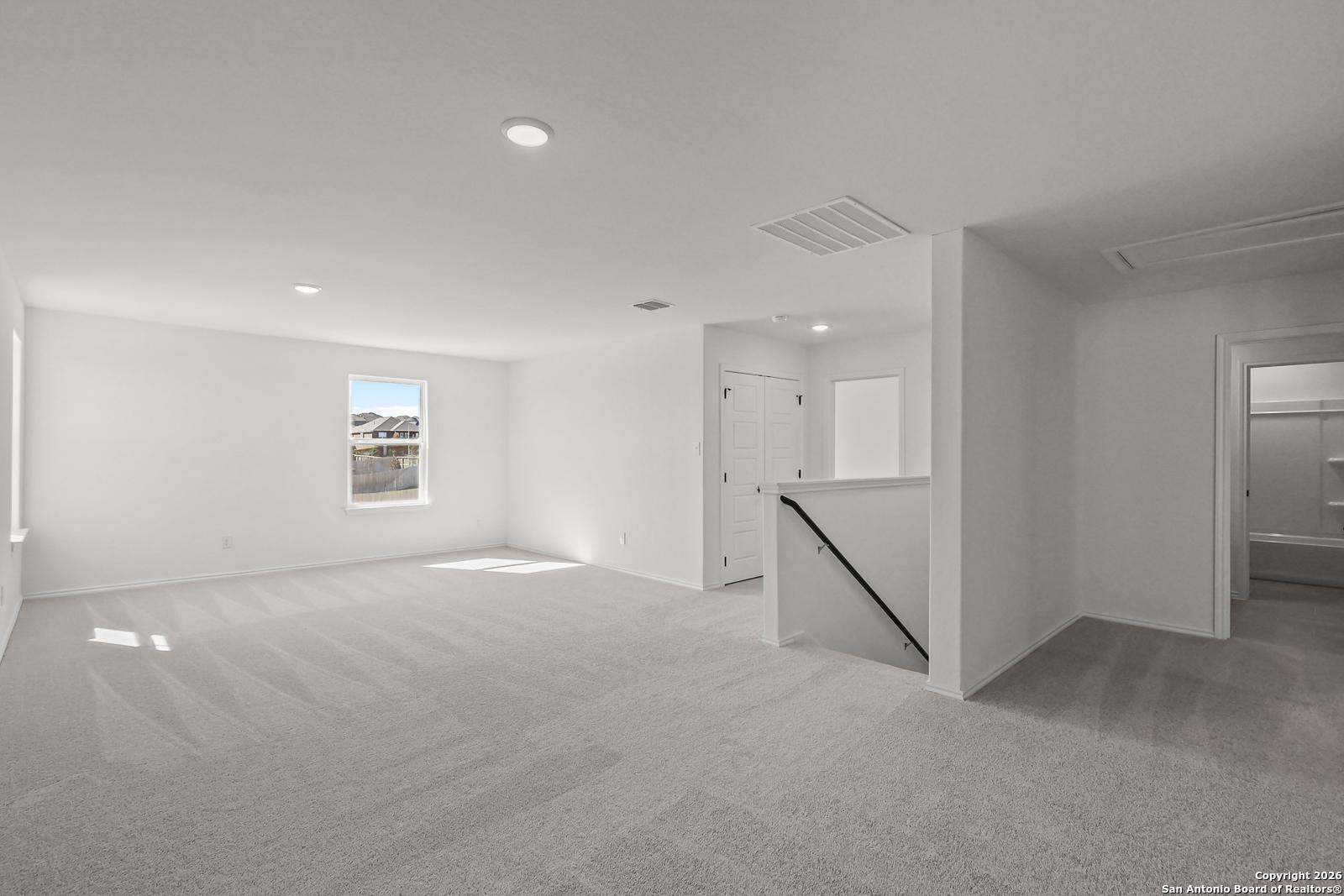 Spacious upstairs loft with white walls, gray carpet, large window, and staircase in Davidson Homes The Douglas E, San Antonio