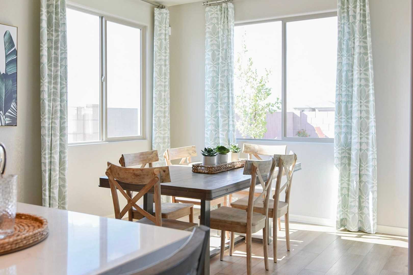 Bright dining area in The Harmony home design featuring wooden table, potted plant, large windows, and adjacent kitchen counter
