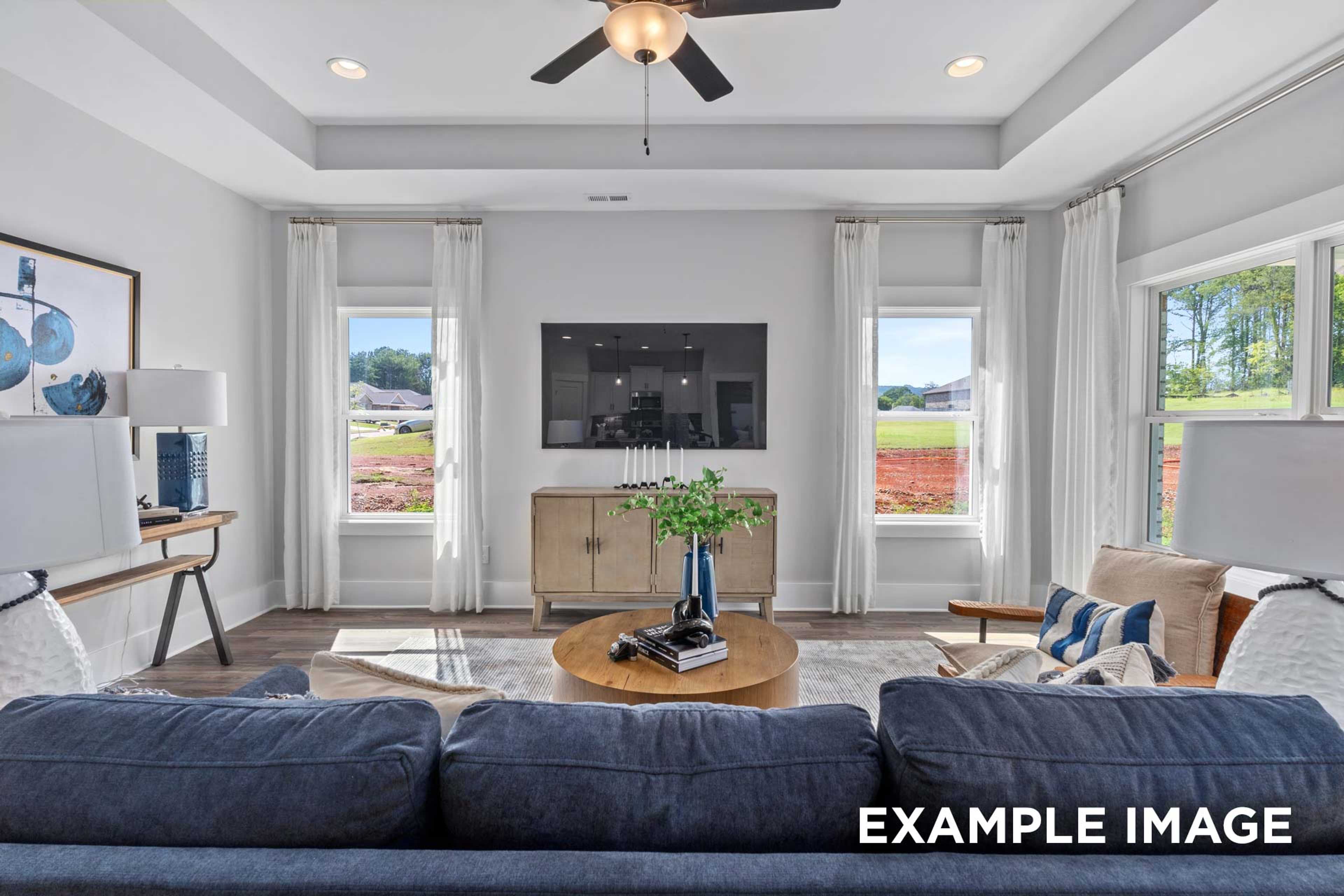Spacious living room in The Franklin C with blue sectional sofa, wall-mounted TV, large windows overlooking greenery, wooden console, ceiling fan