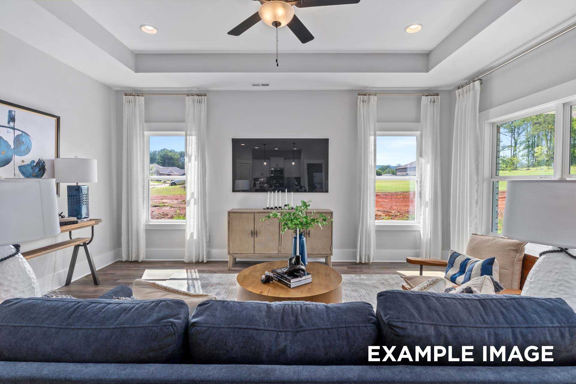 Spacious living room in The Franklin C with blue sectional sofa, wall-mounted TV, large windows overlooking greenery, wooden console, ceiling fan
