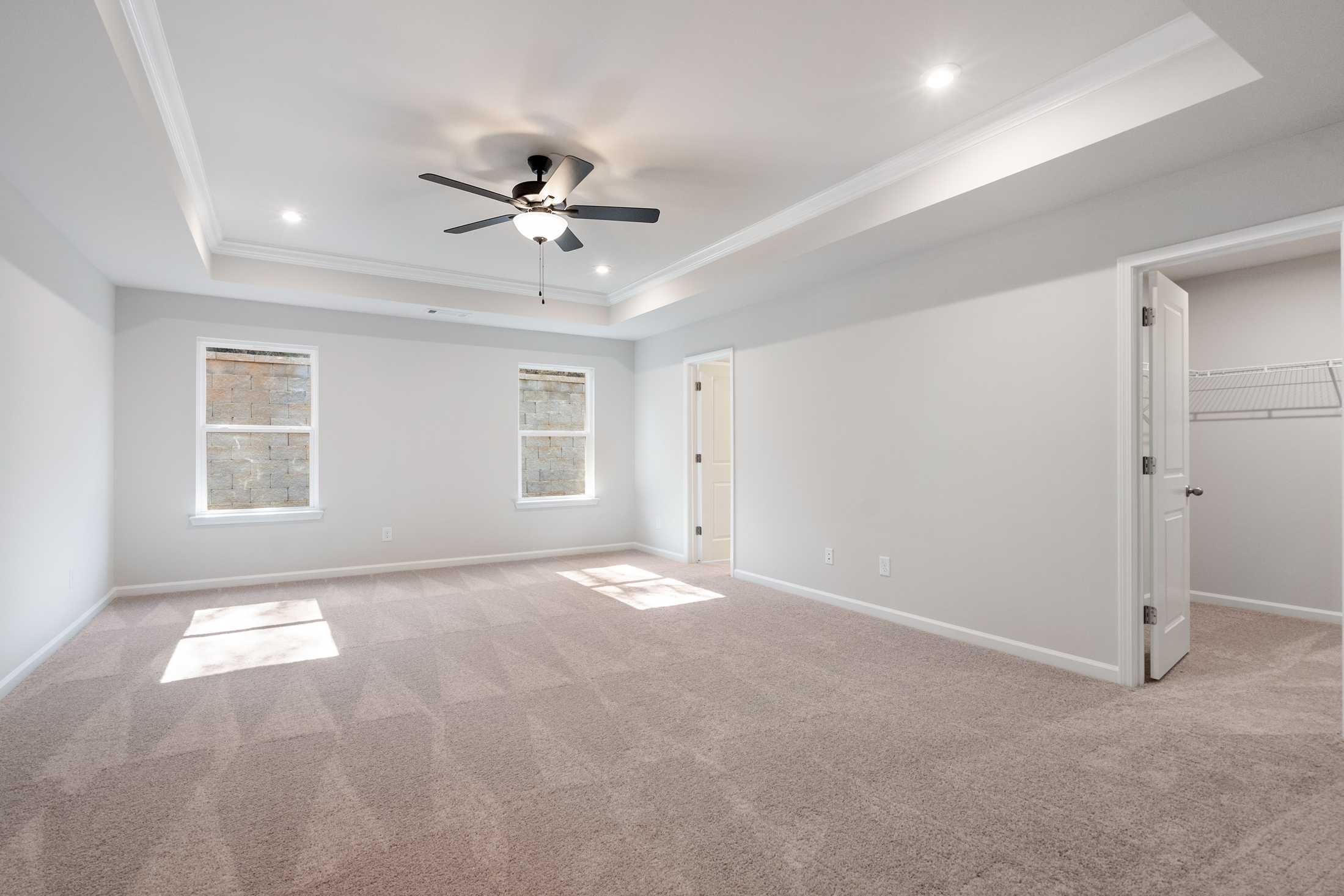 Spacious master bedroom in The Cary B featuring beige carpet, ceiling fan, tray ceiling, large windows, and walk-in closet