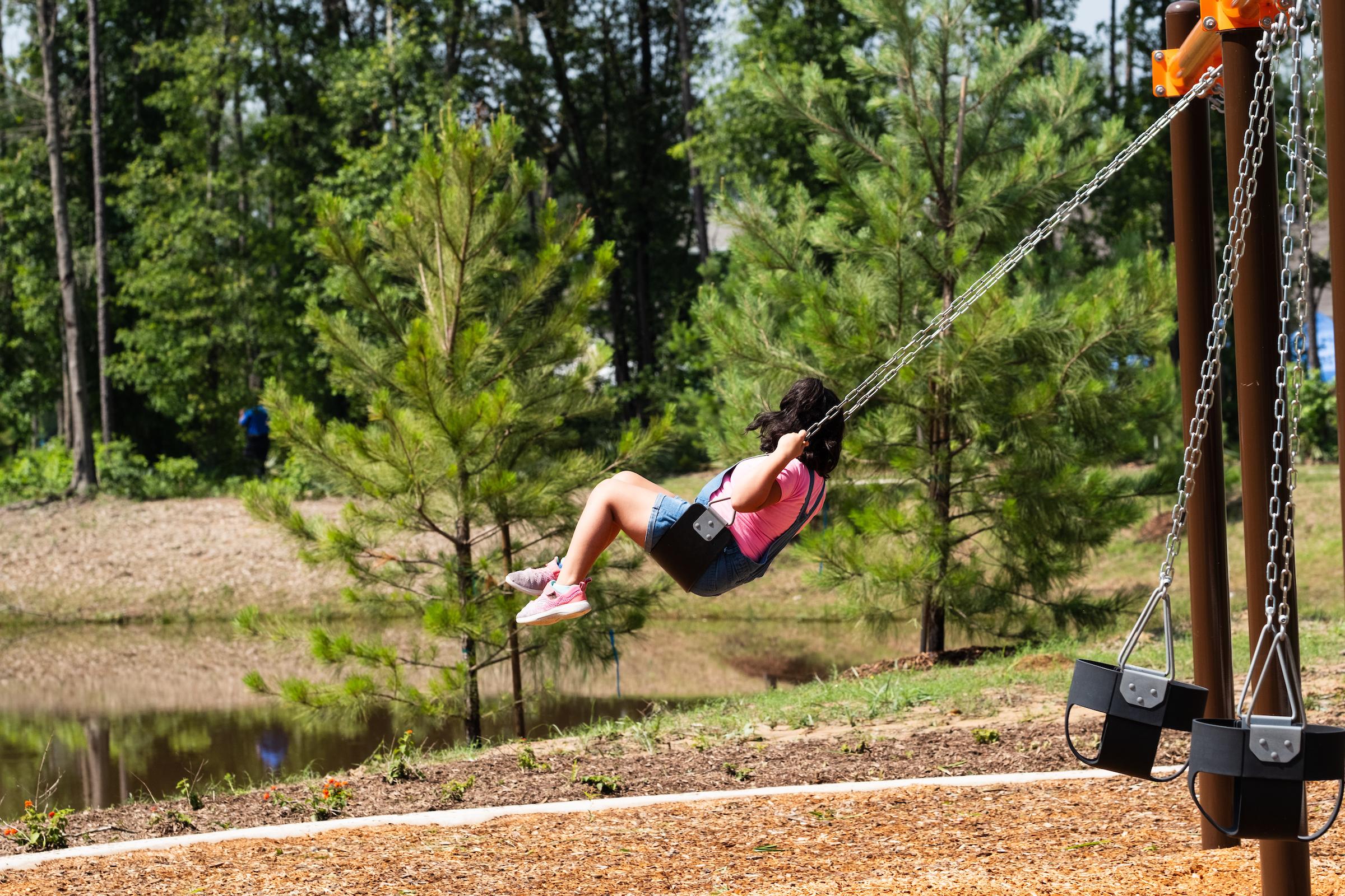 Child swinging on playground set at Robins Landing in Houston Texas amid pine trees and scenic pond