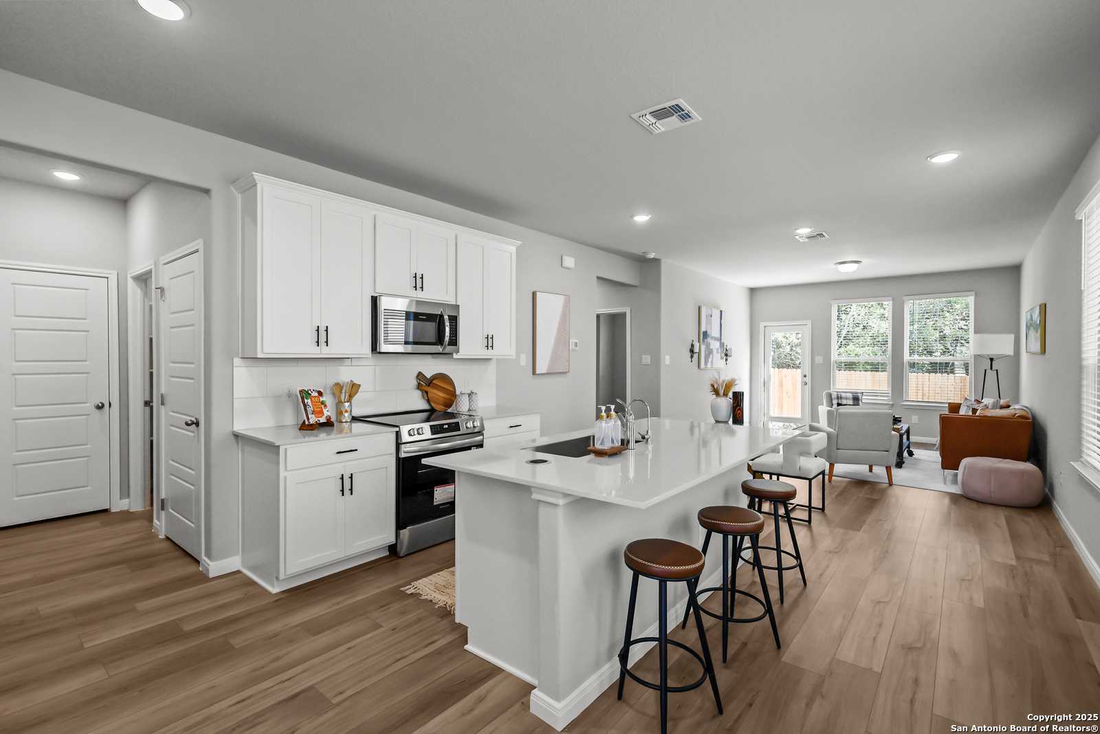 Modern open kitchen with white cabinets, stainless appliances, island bar stools, and hardwood floors in The Gillian C, San Antonio home