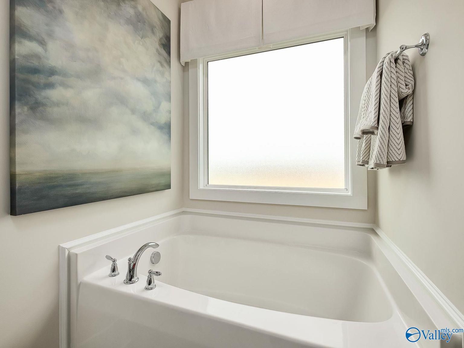 Elegant master bathroom featuring white corner soaking tub, large window with sheer curtains, abstract seascape art in Davidson Homes The Everett, Toney, AL