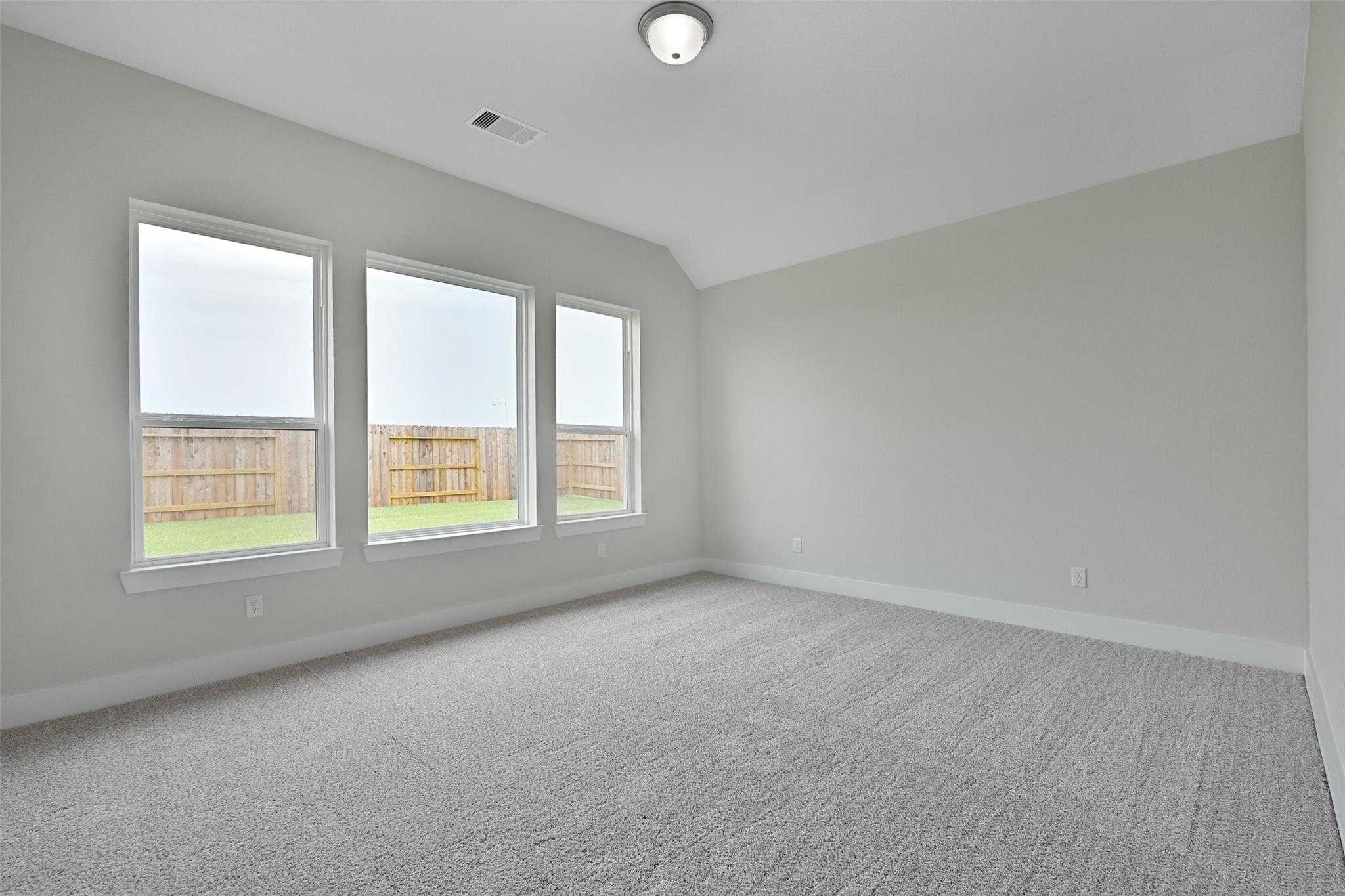 Bright empty bedroom with three large windows overlooking fenced backyard, gray walls, beige carpet in Davidson Homes Edward C, Texas City