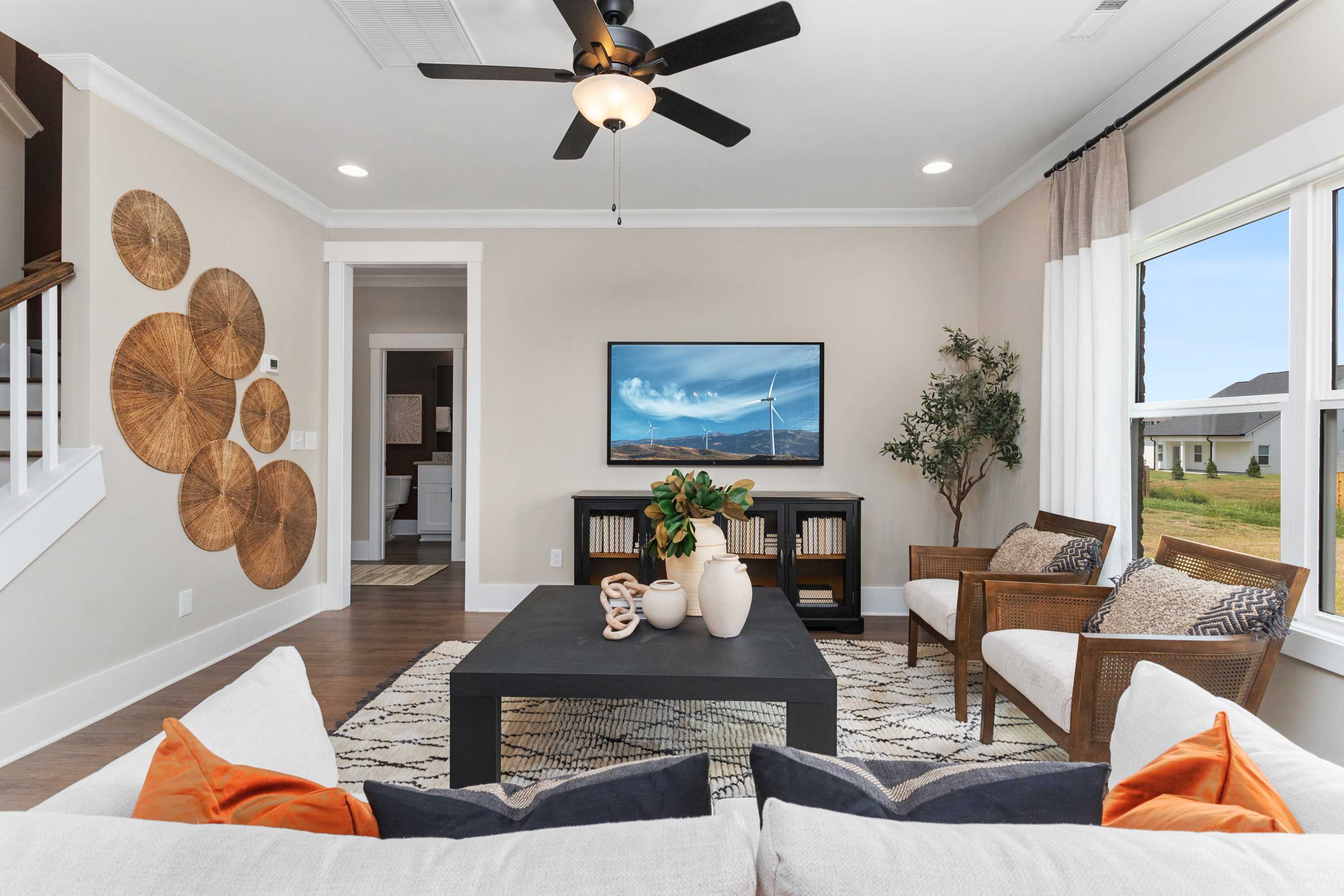 Cozy living room in The Shelby A by Davidson Homes, with ceiling fan, woven wall art, TV, large windows, and modern seating
