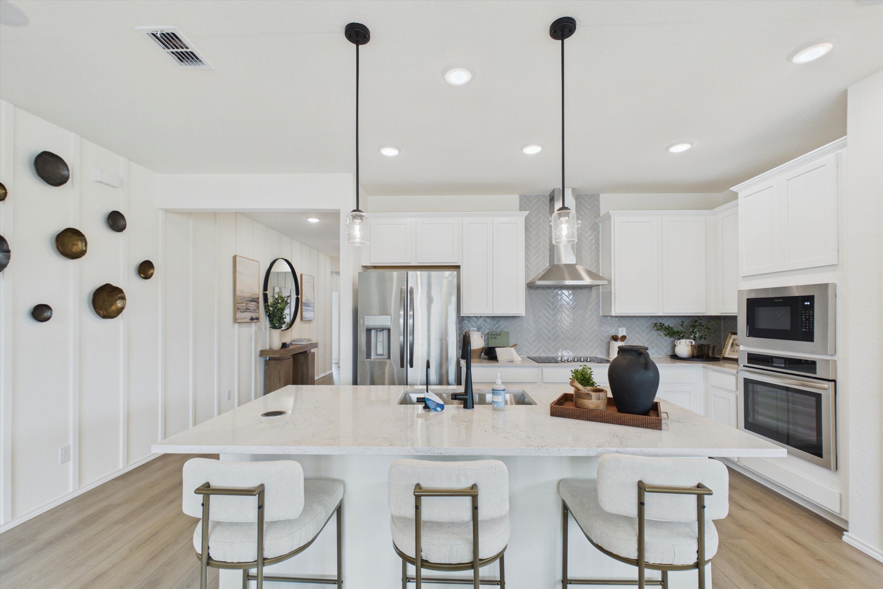 Spacious modern kitchen in Rayburn Valley San Antonio TX by Davidson Homes with white shaker cabinets, quartz island, herringbone backsplash, stainless appliances, and upholstered bar stools