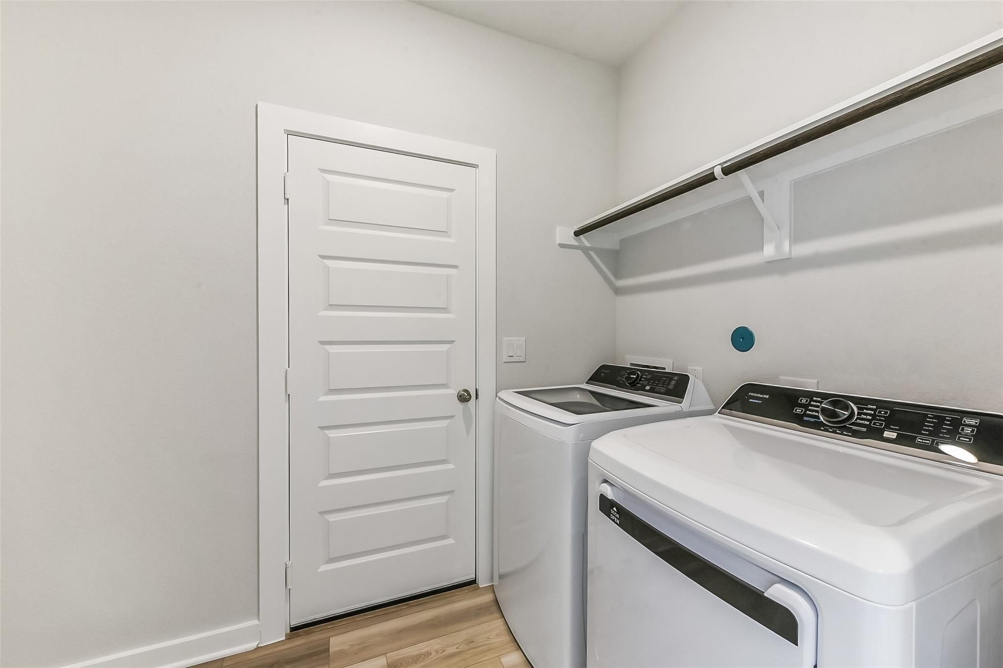 Modern laundry room featuring white Frigidaire washer-dryer set, overhead shelves, and wood flooring in Davidson Homes The Frio G, Magnolia TX