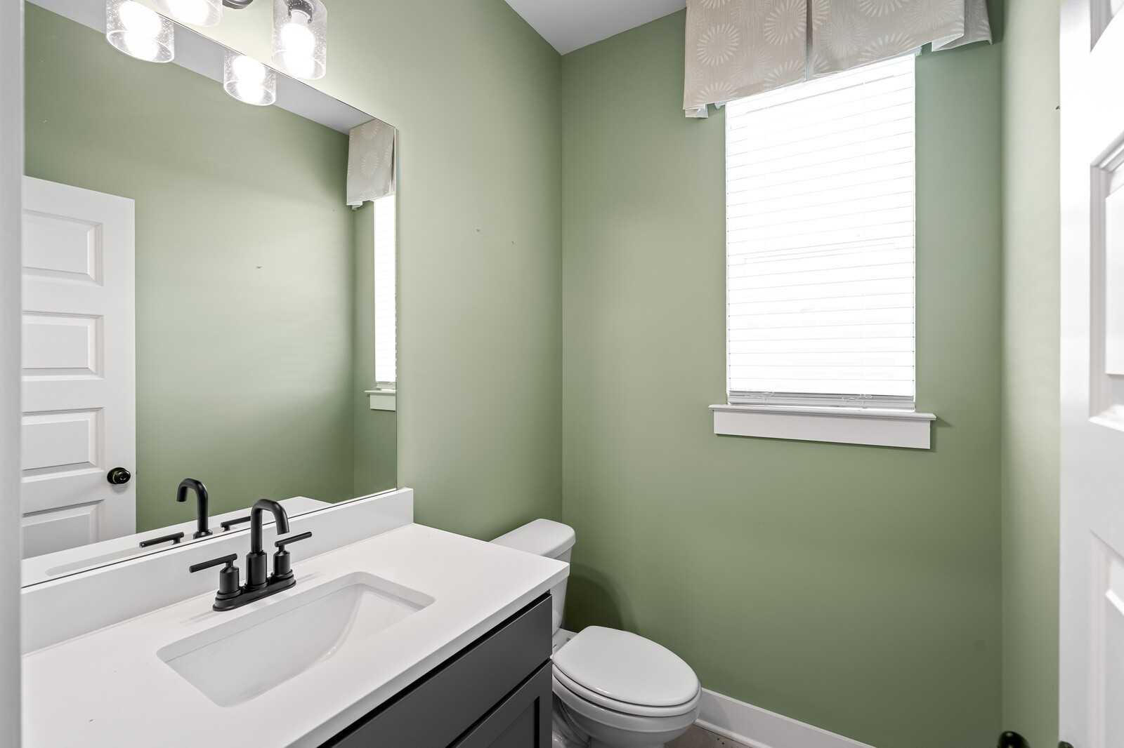 Sage green powder room with white vanity, black faucet, large mirror, toilet, and window blinds in Davidson Homes The Ridgeport, Gallatin, TN