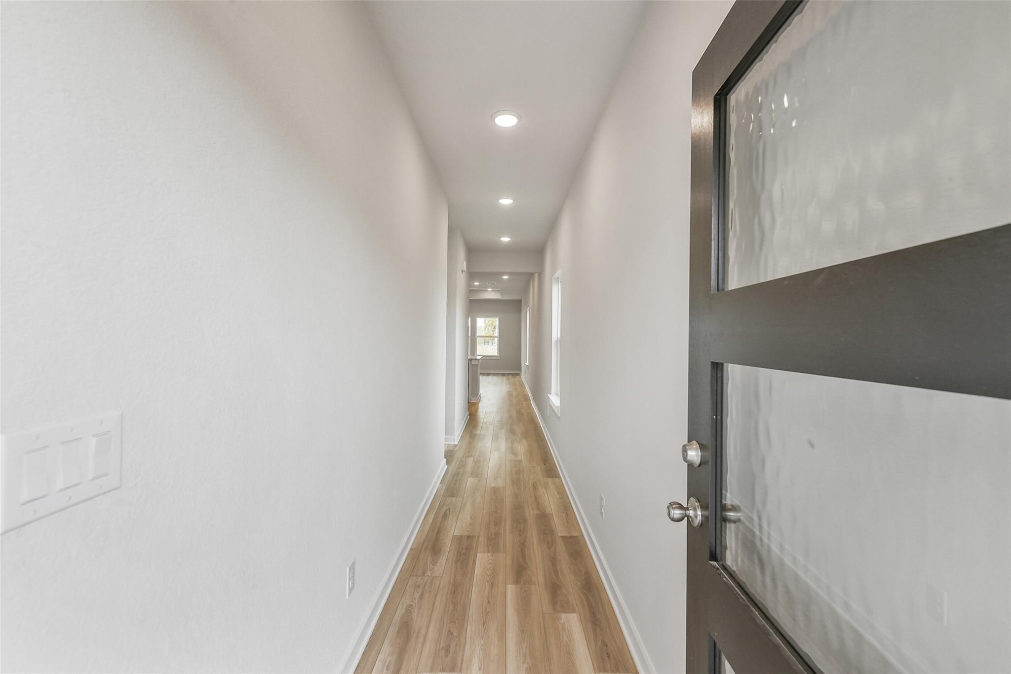 Long hallway with light oak hardwood floors, white walls, recessed lighting, and frosted glass front door in Davidson Homes Colorado G, Magnolia TX