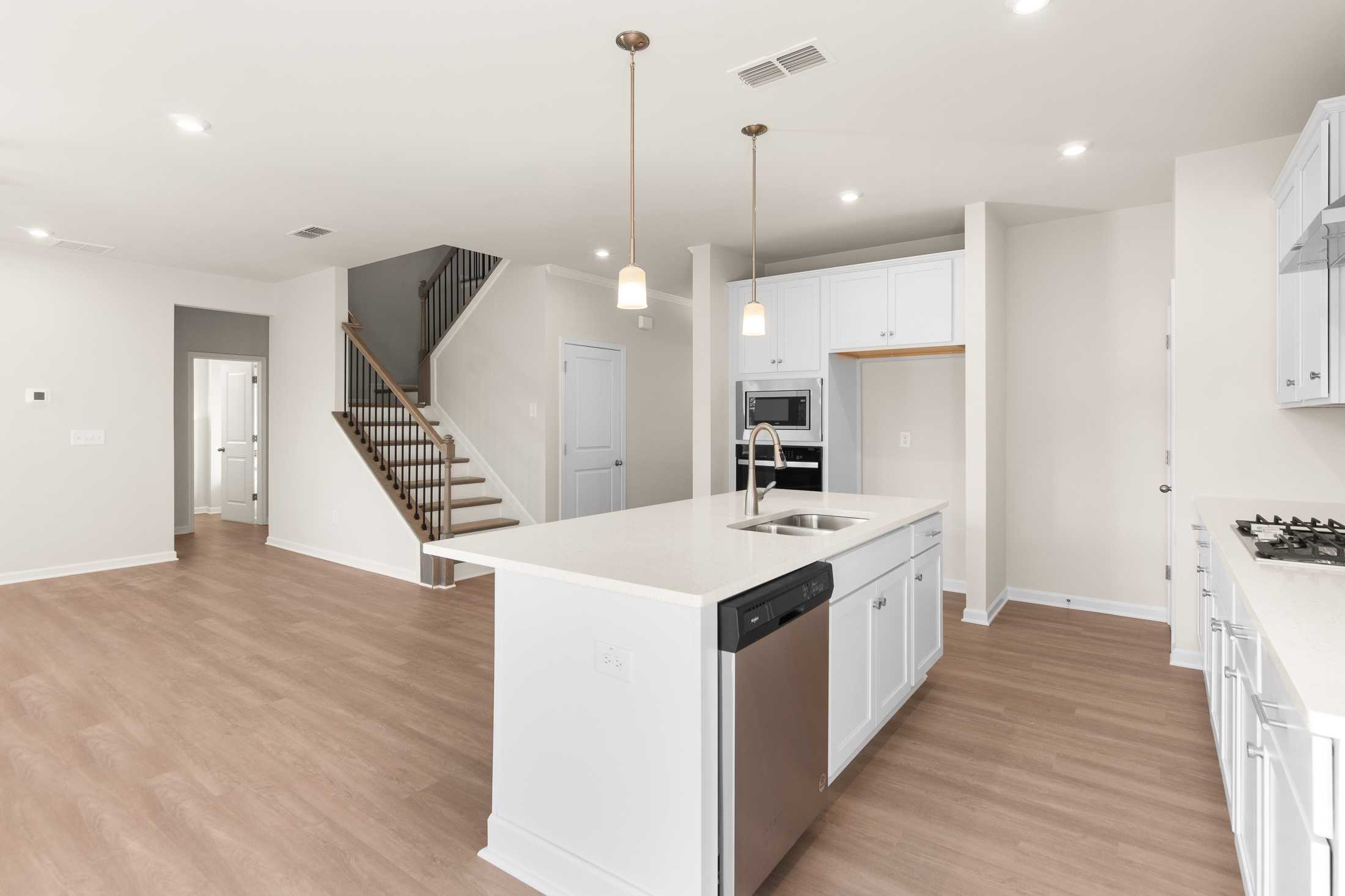 Spacious open-concept kitchen in The Willow B with white island, sink, dishwasher, wood floors, and staircase