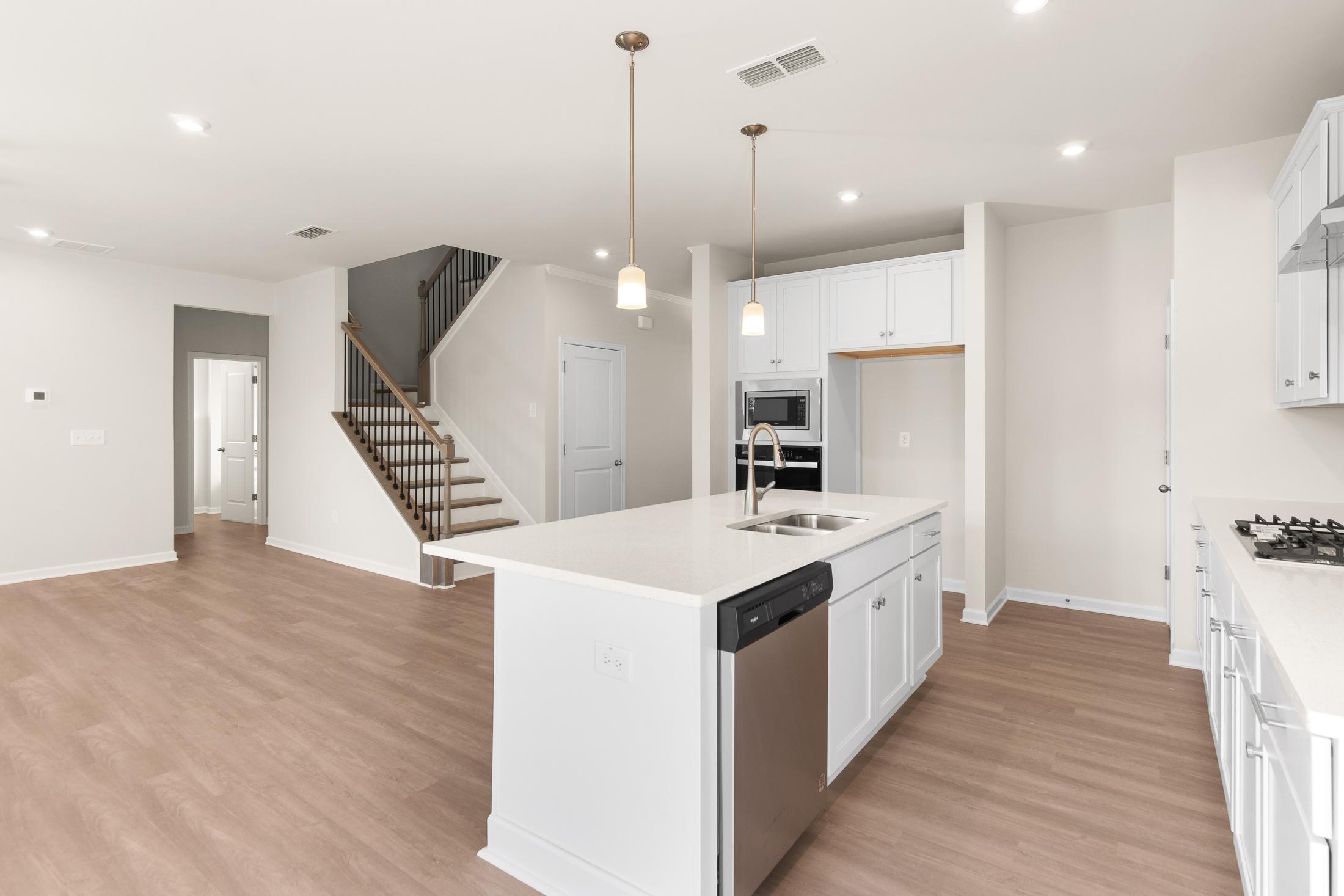Spacious open-concept kitchen in The Willow B with white island, sink, dishwasher, wood floors, and staircase