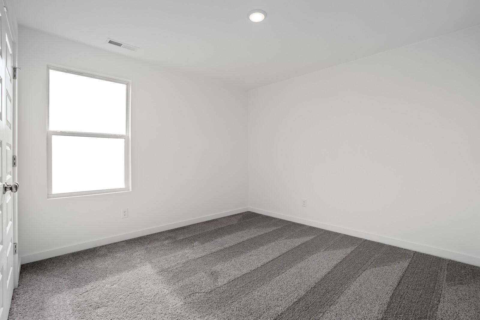 Bright empty bedroom featuring large window, white walls, and gray carpet in Davidson Homes The Logan C, White House, Tennessee