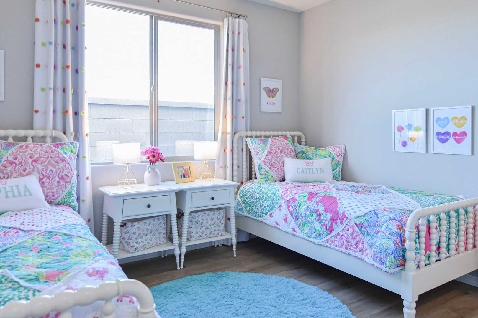 Cozy girls' twin bedroom in The Harmony with floral pink-green quilts, white nightstands, polka dot curtains, and butterfly wall art