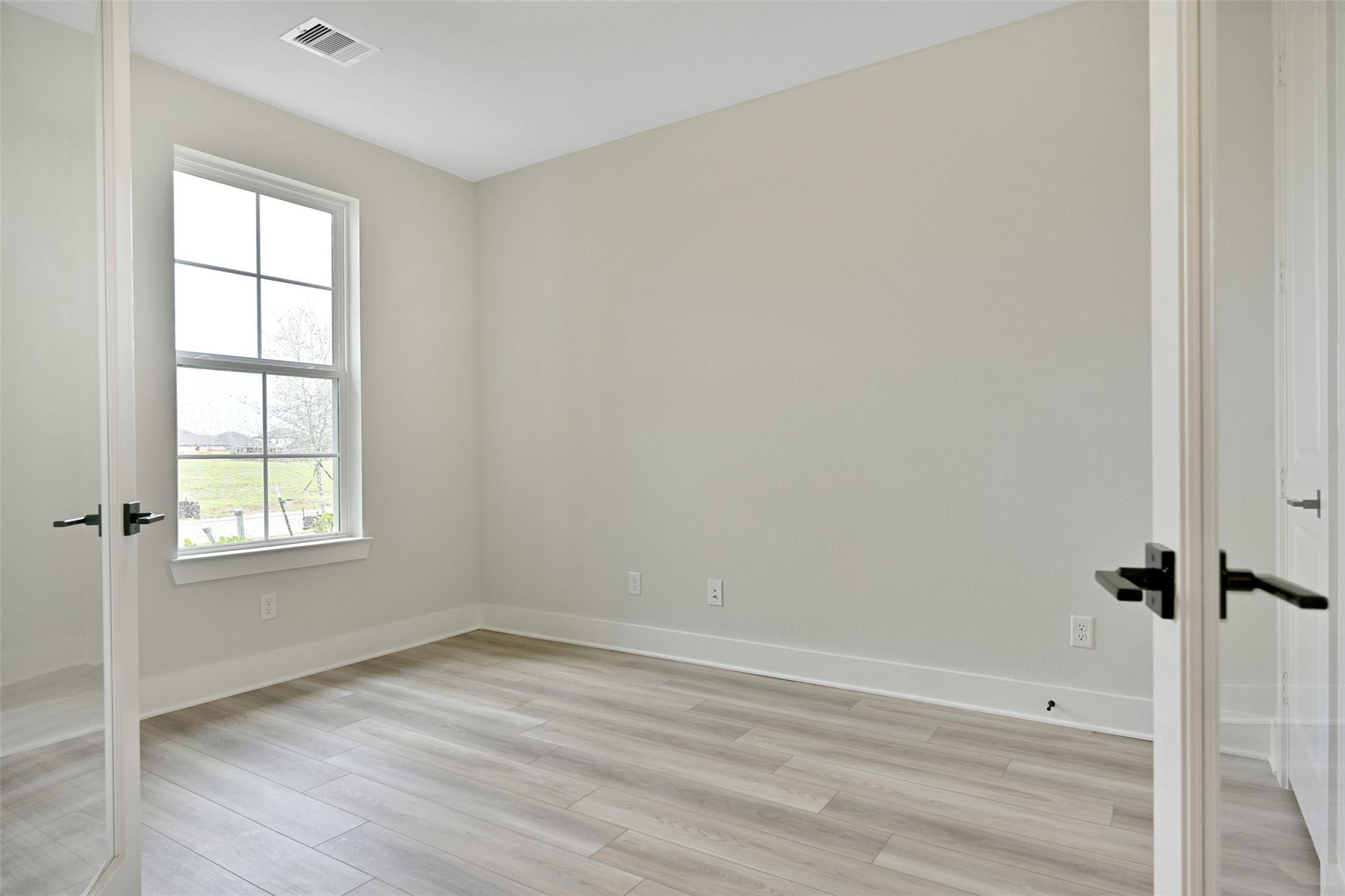 Empty bedroom with light laminate flooring, beige walls, large window, and open doors in Davidson Homes The Edward C, Lago Mar, Texas City