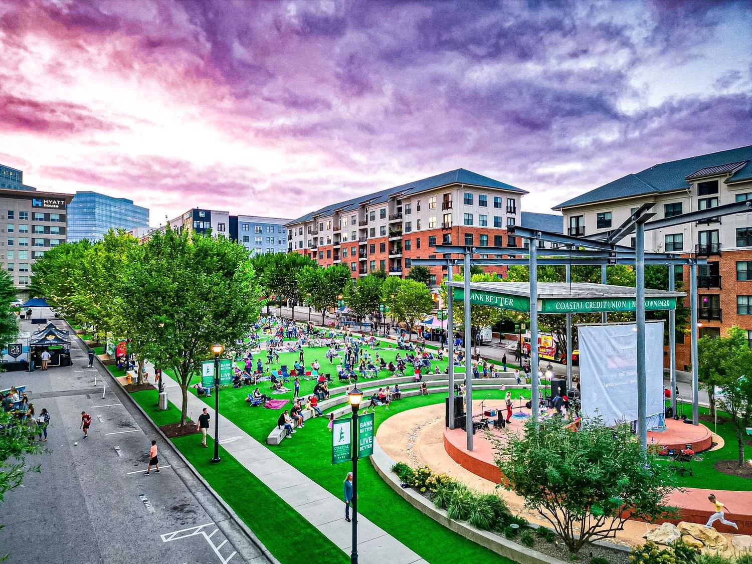 Outdoor community event at Coastal Credit Union Midtown Park in Wake Forest, NC, featuring a stage and green lawn.