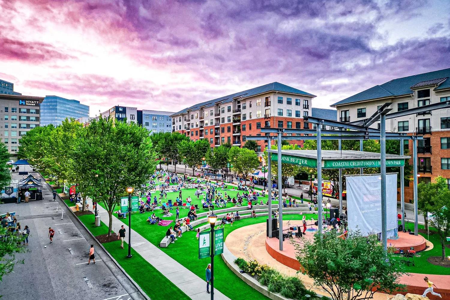 Outdoor community event at Coastal Credit Union Midtown Park in Wake Forest, NC, featuring a stage and green lawn.
