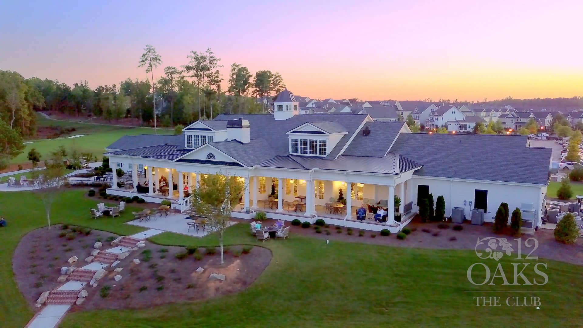 Aerial view of elegant clubhouse at Addison West, Holly Springs NC, with white verandas, oak trees, and sunset over golf course