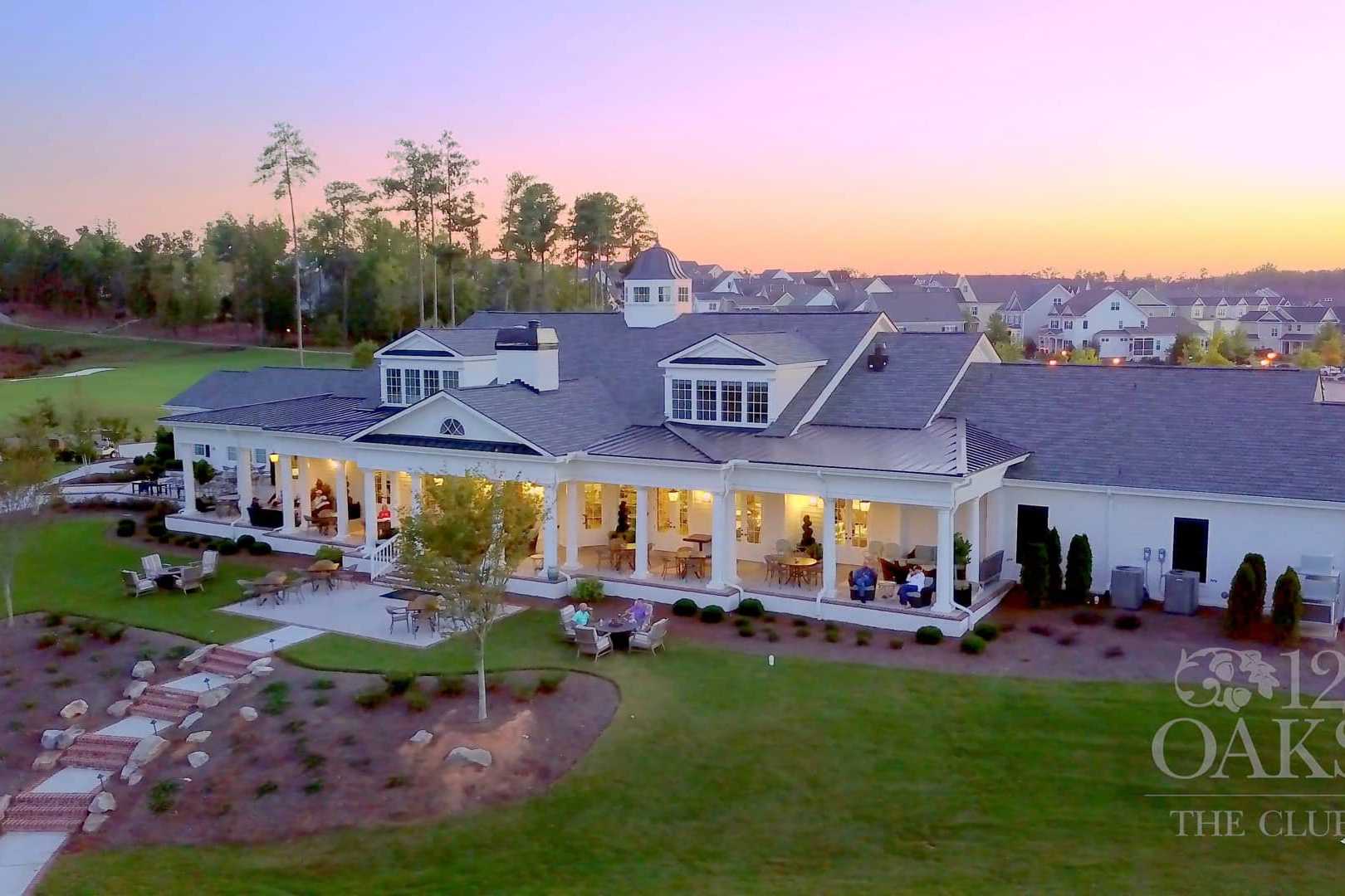 Aerial view of elegant clubhouse at Addison West, Holly Springs NC, with white verandas, oak trees, and sunset over golf course
