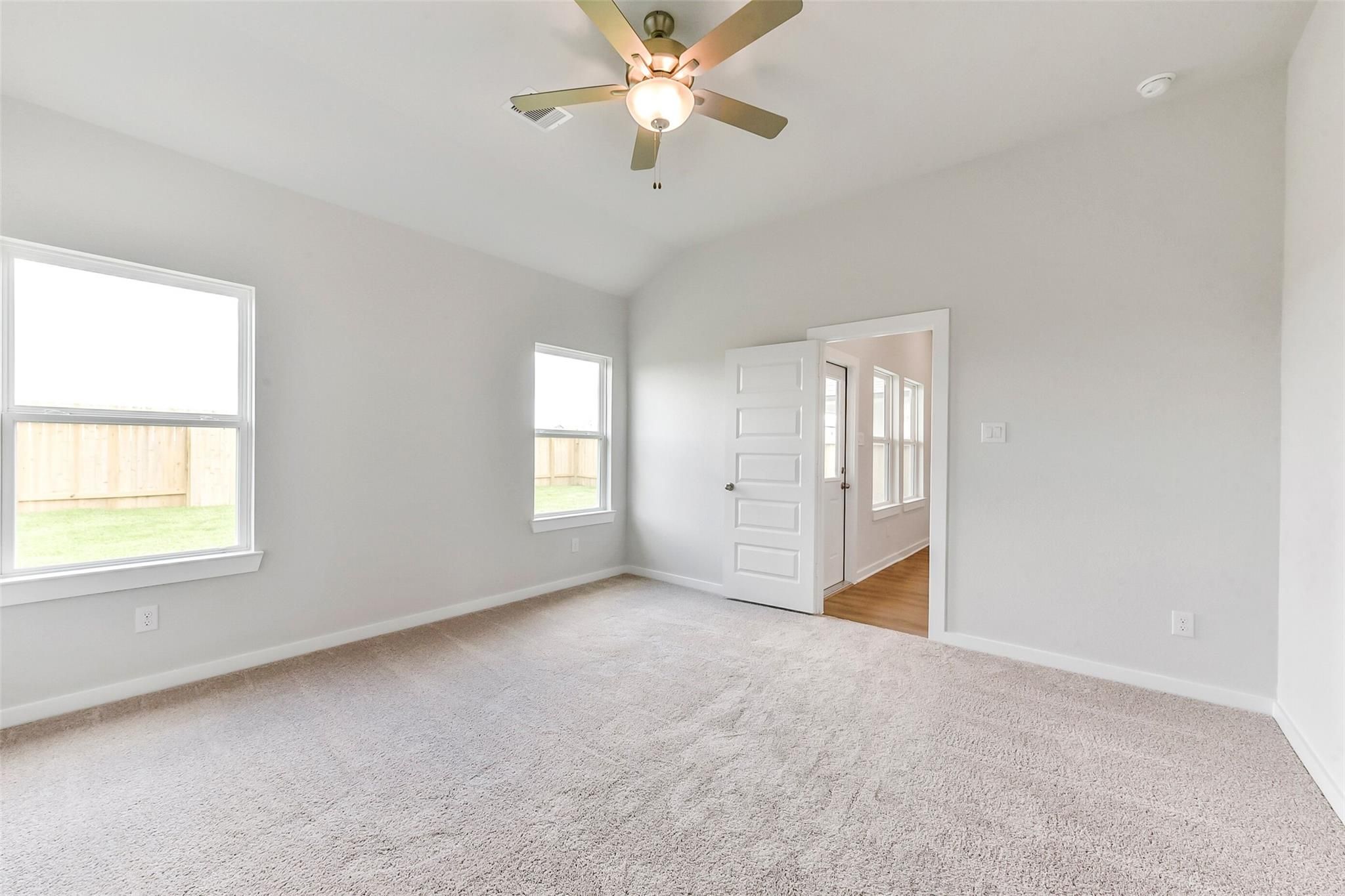 Bright bedroom with beige carpet, ceiling fan, large windows, and open door in Davidson Homes The Costa B, Dayton, Texas