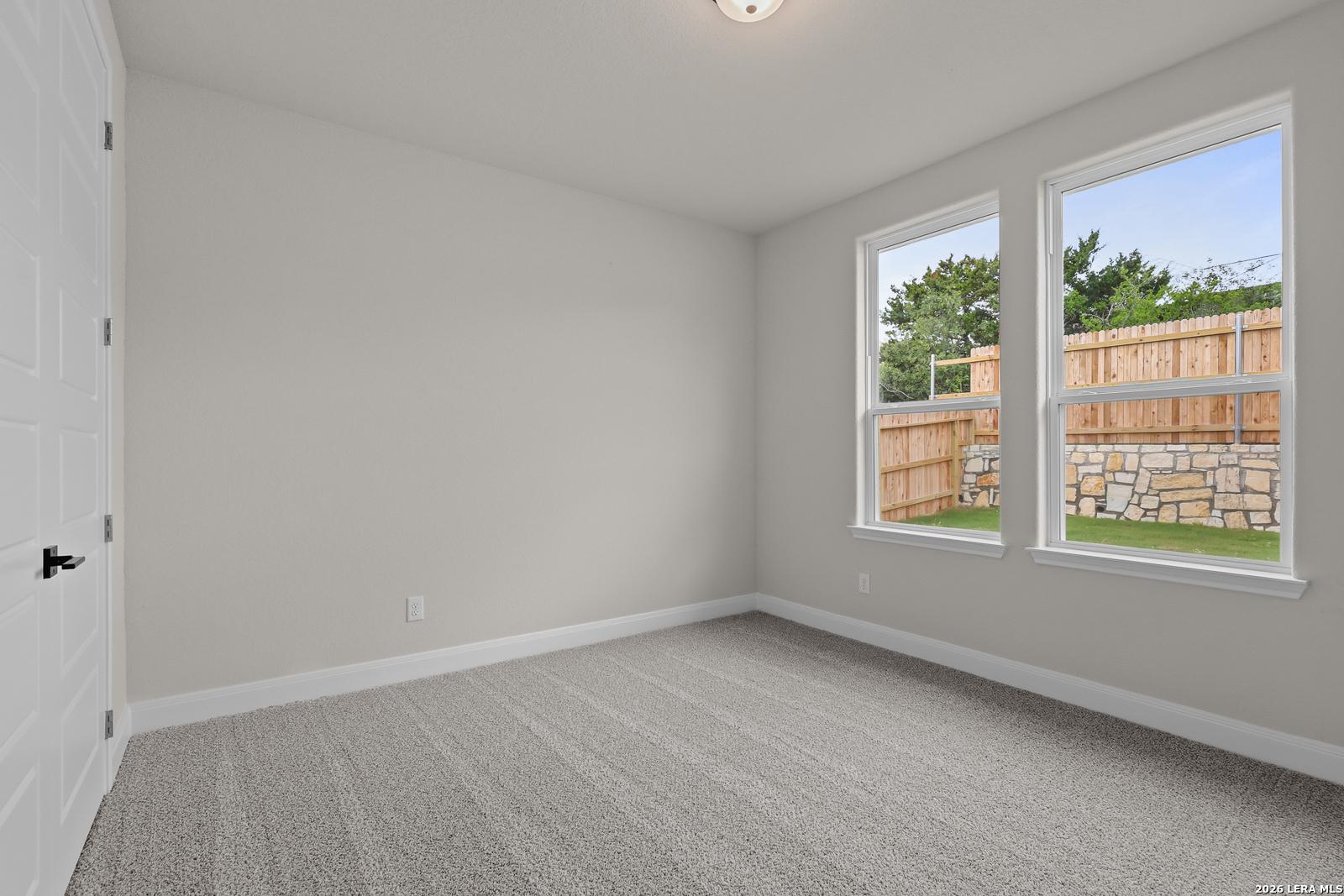 Bright secondary bedroom with gray carpet, light walls, and large windows overlooking fenced backyard in The Lanier G, Castroville, TX
