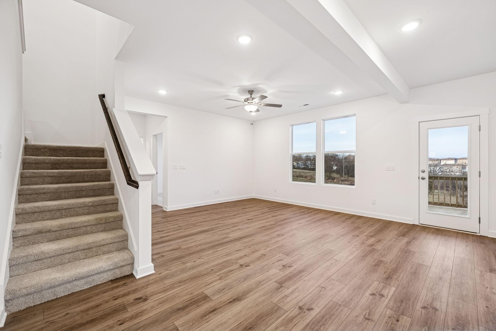 Spacious open living room with hardwood floors, carpeted staircase, ceiling fan, and large windows to countryside in The Willow home, Calista Farms
