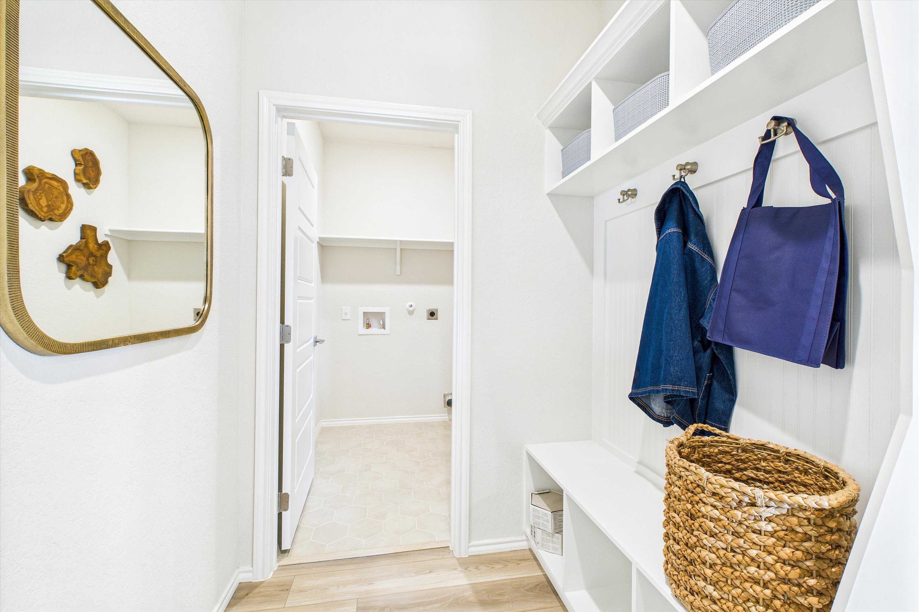 Spacious mudroom at Fairway Crossing in Converse Texas with built-in white cabinetry, coat hooks, bench, wicker basket, and adjacent laundry room