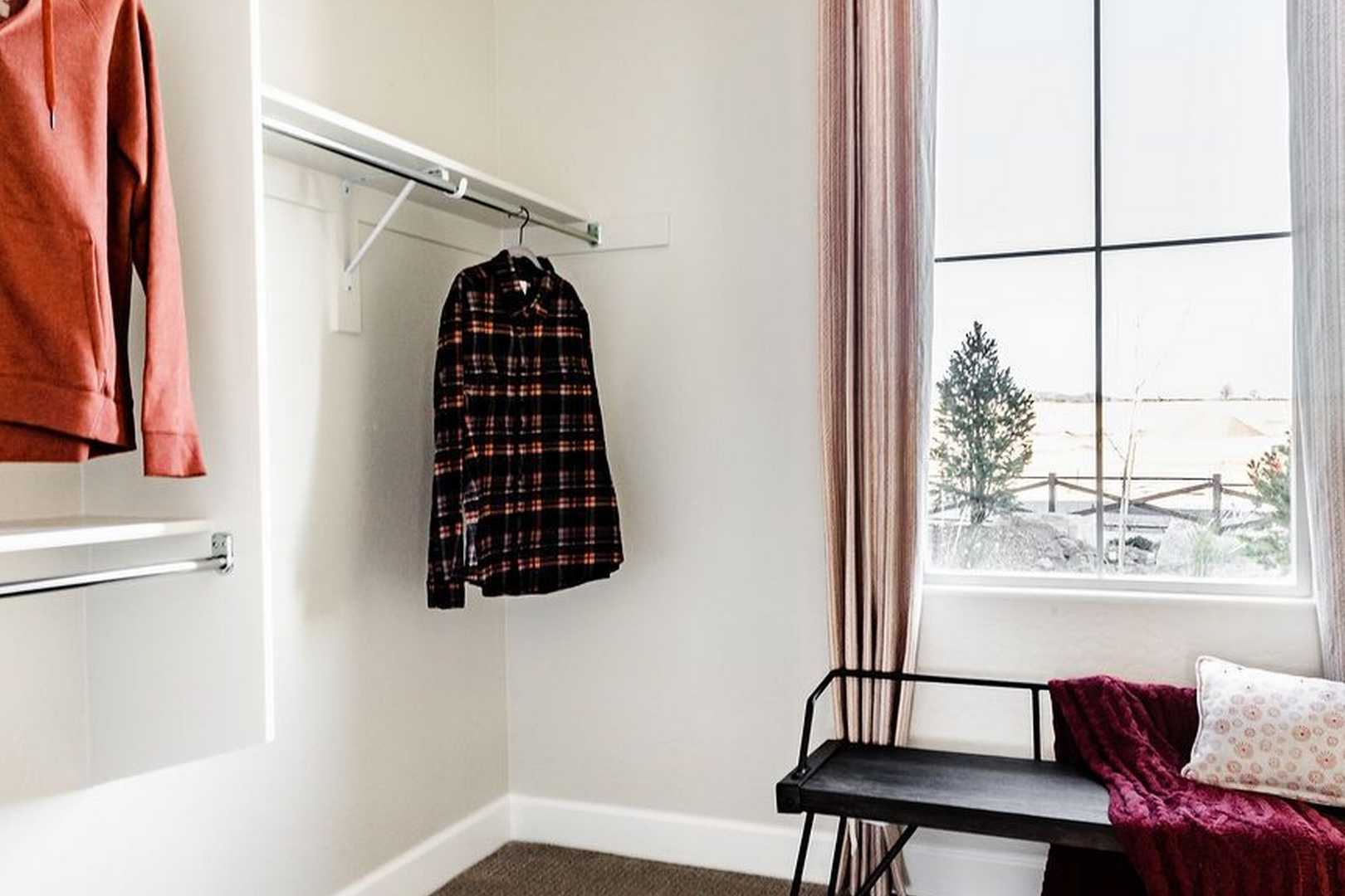 Spacious master suite closet in The Monarch featuring hanging plaid shirt and hoodie, built-in bench with throw, window view