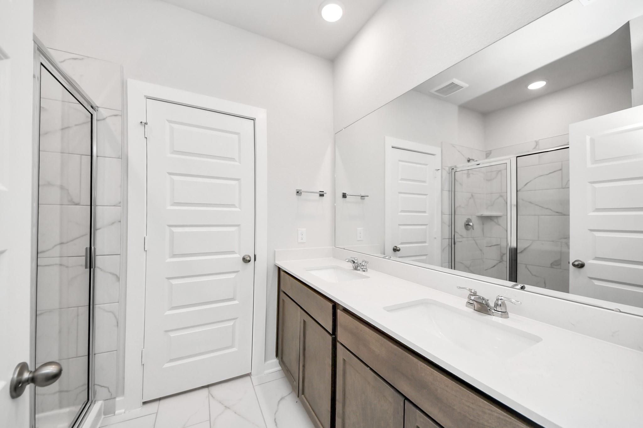 Modern primary bathroom with dual vanity, large mirror, and glass-enclosed shower in The Sabine E at Liberty Estates.