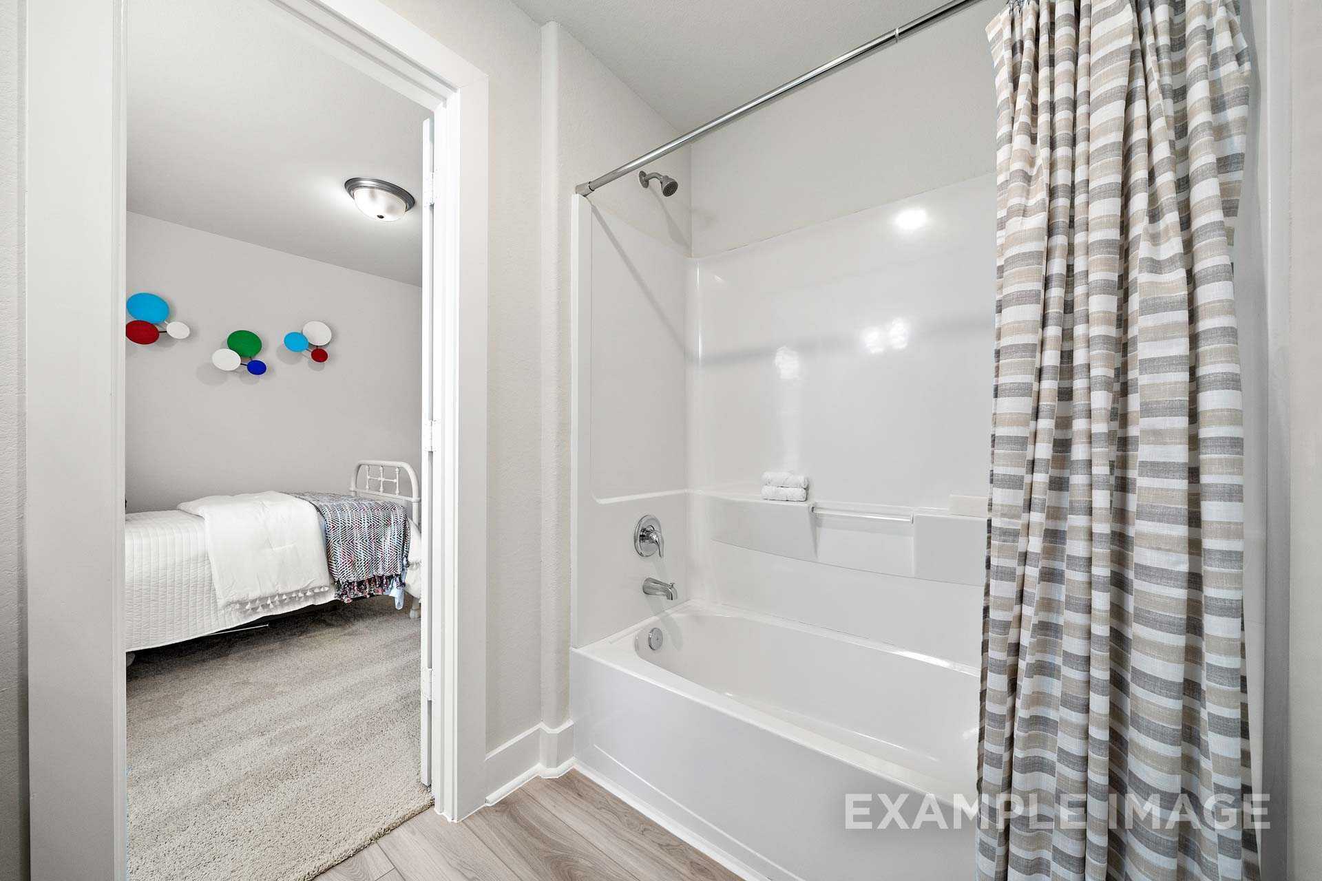 The Lily A master bathroom with white tub, walk-in shower, gray curtain, open to cozy bedroom with colorful wall art