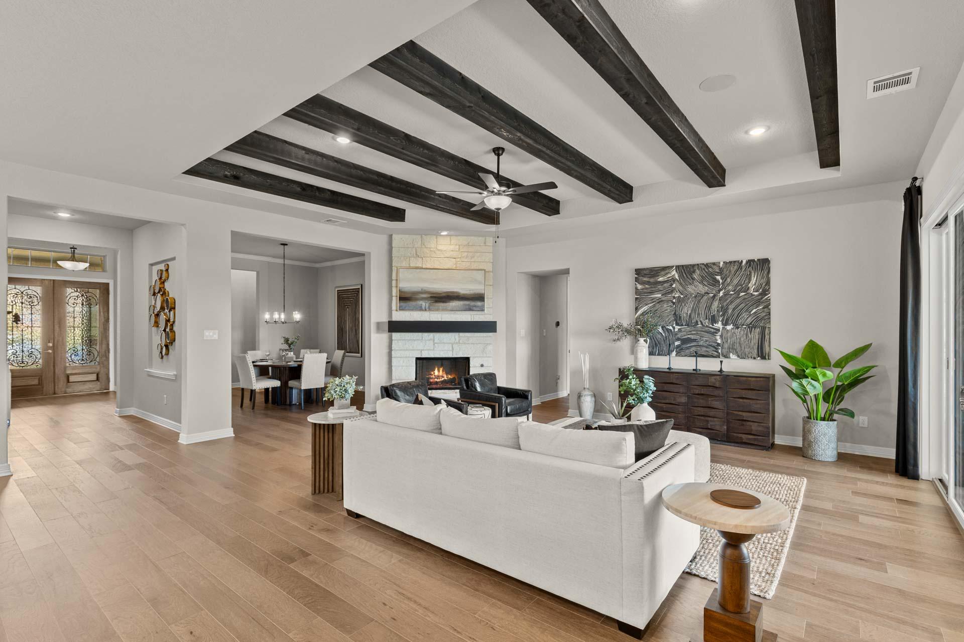 Open-concept great room at Potranco Oaks in Castroville TX by Davidson Homes featuring beamed ceiling, hardwood floors, white sofa, and stone fireplace