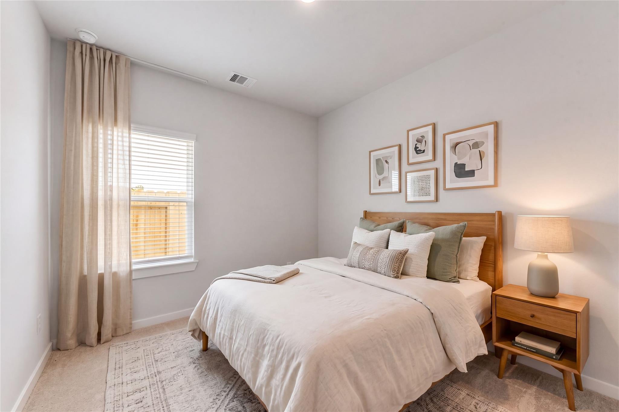 Serene bedroom with wooden bed, neutral linens, green pillows, abstract wall art, and window in Davidson Homes The Sabine F, Dayton, Texas
