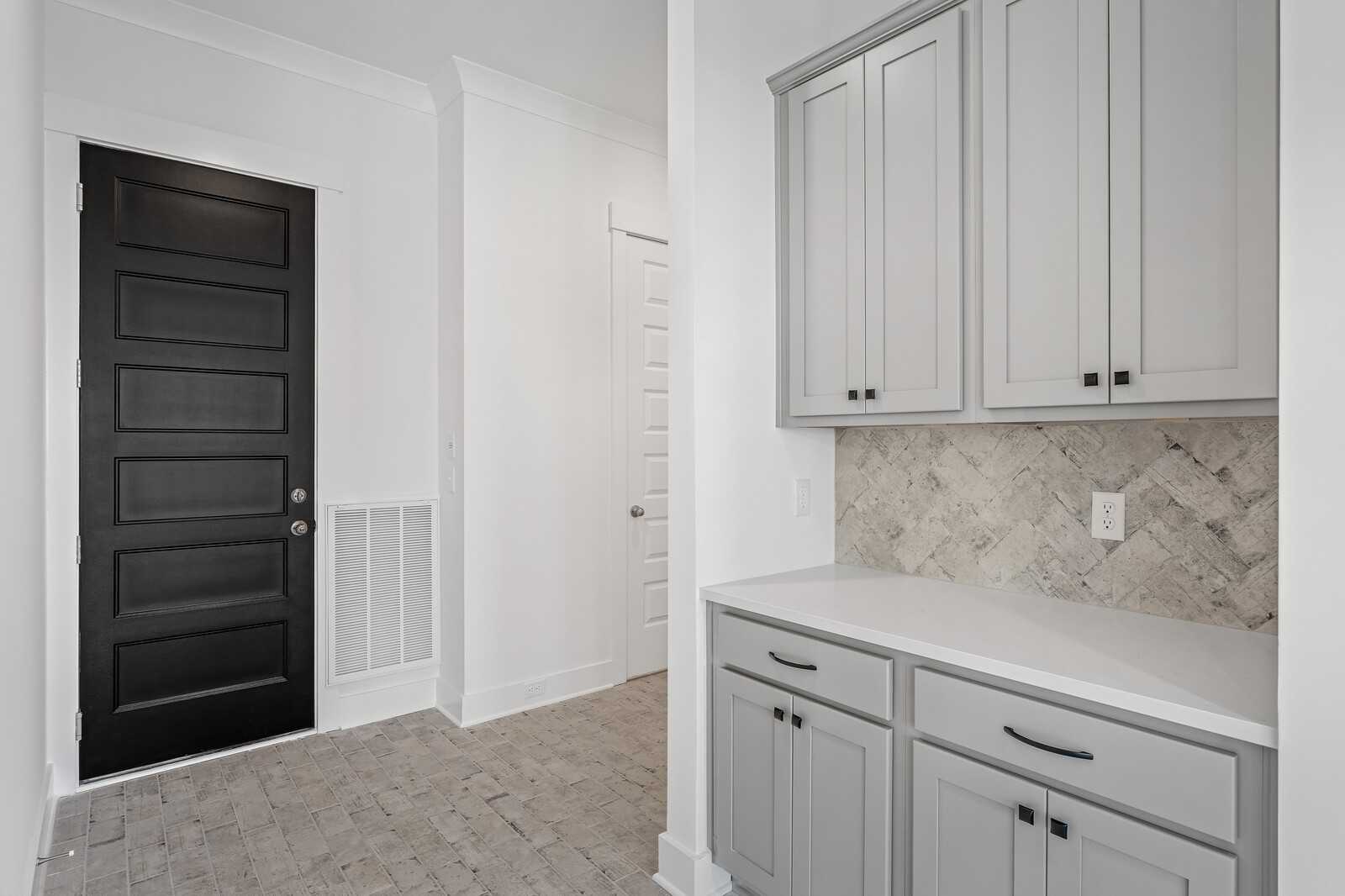 Modern laundry room with gray shaker cabinets, white quartz counter, herringbone tile backsplash, and black door in The Alston A, Murfreesboro