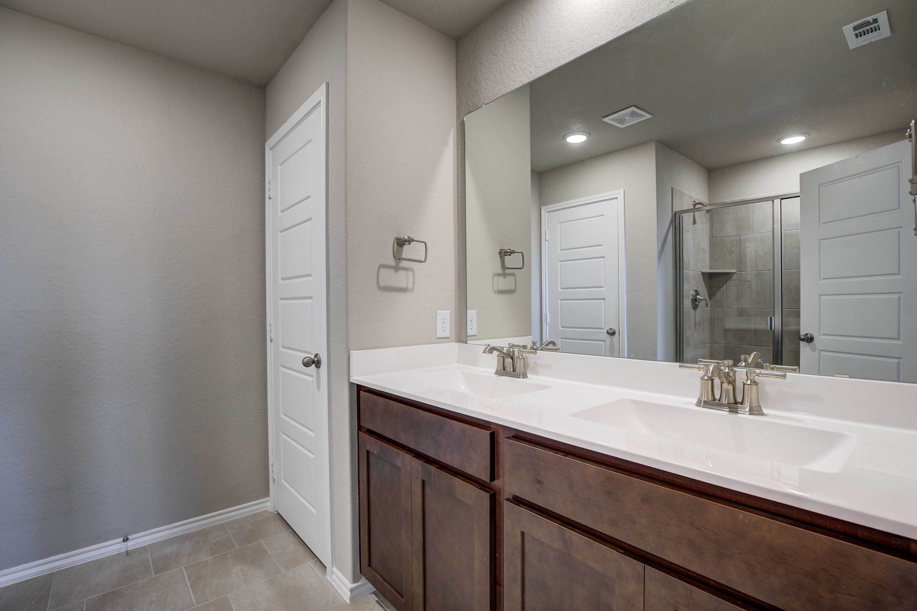 Spacious master bathroom in The Murray with double vanity, brown cabinets, large mirrors, and glass shower door