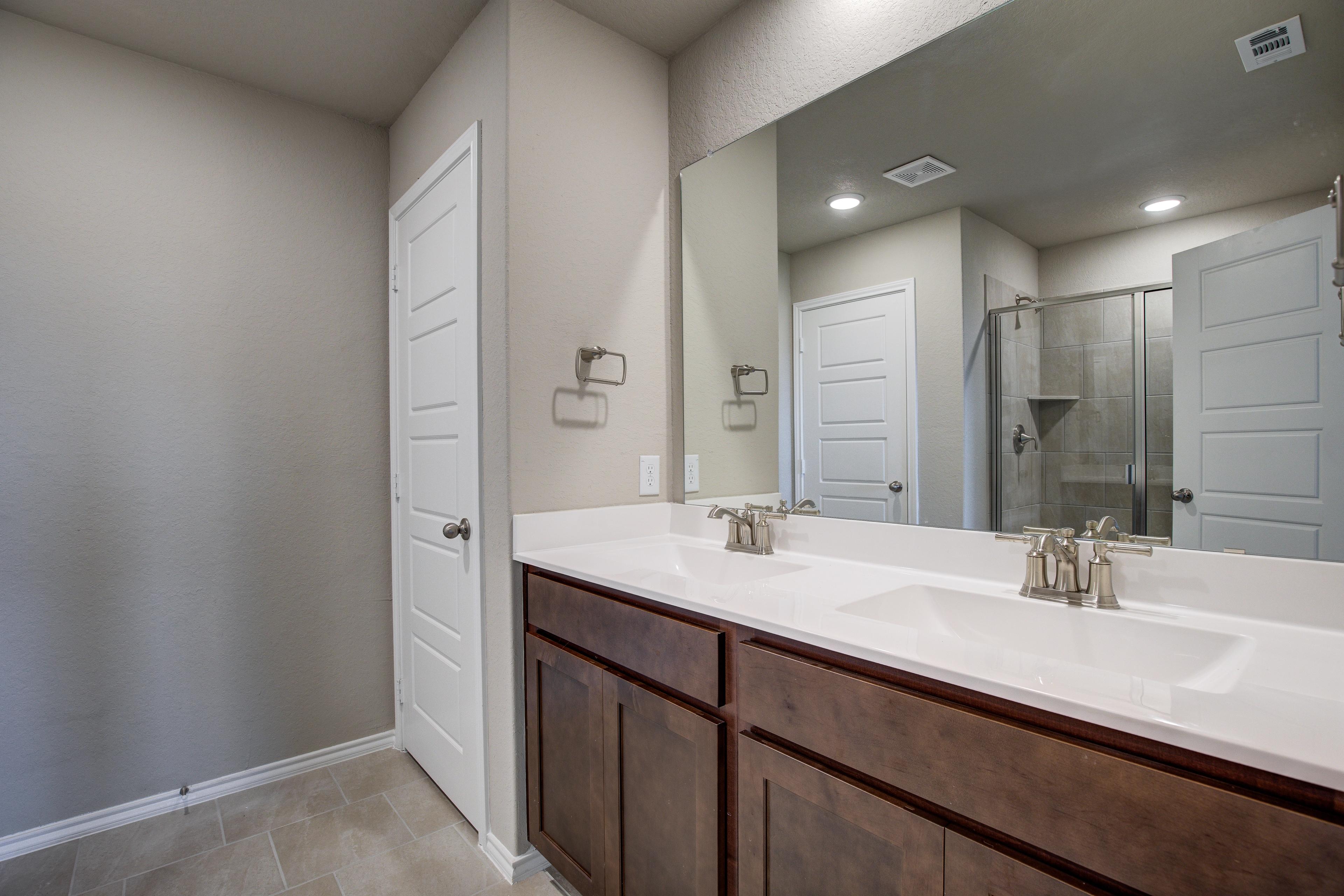 The Murray G master bathroom with double sink vanity, brown cabinets, large mirror, and glass shower door in San Antonio home