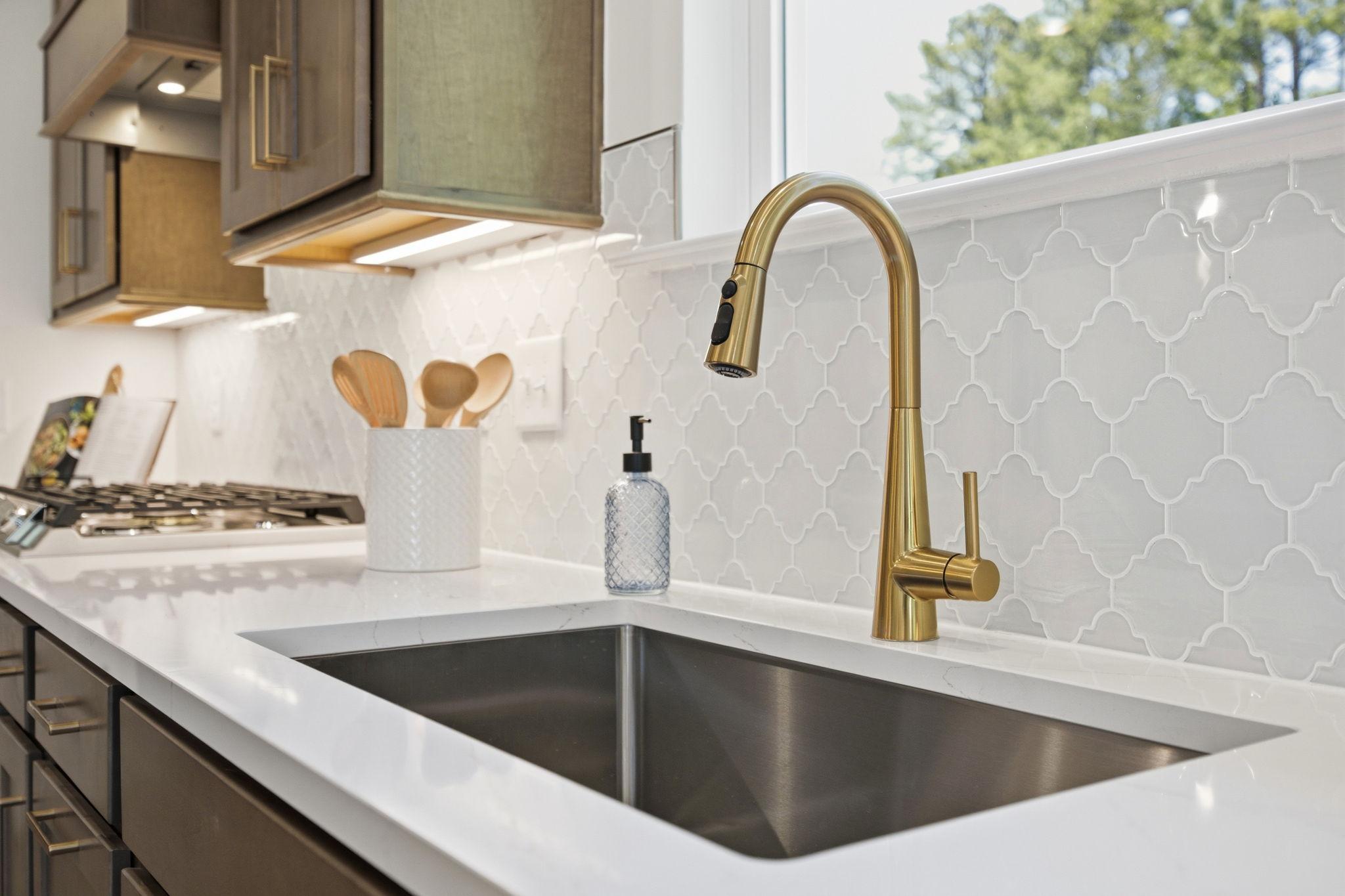 Modern kitchen design in The Ashport featuring a gold faucet, stainless steel sink, and white arabesque tile backsplash.