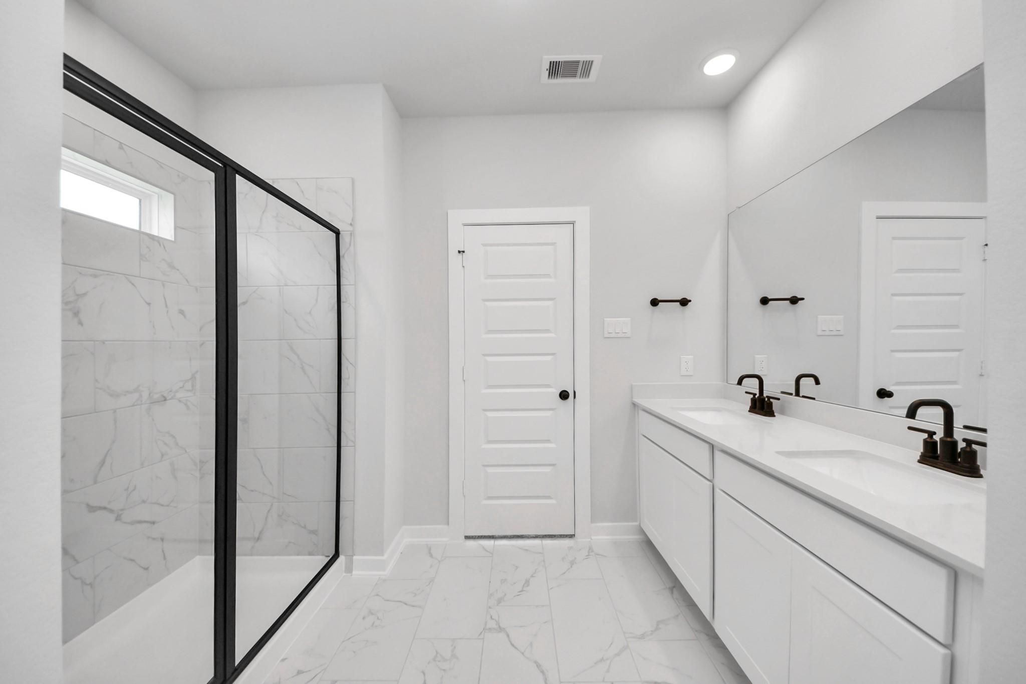 Modern primary bathroom with double vanity, walk-in tiled shower, and black fixtures in The Everett C, Dayton, Texas home.