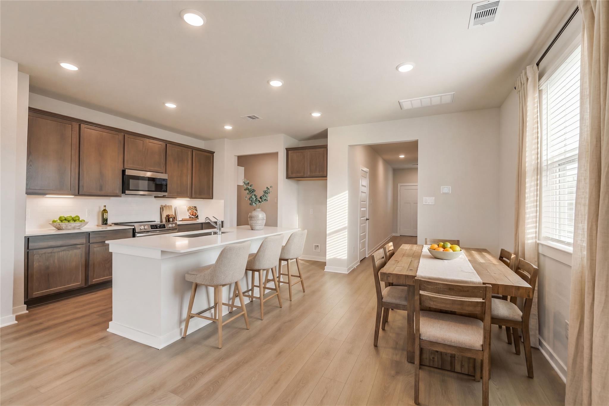 Modern open-concept kitchen with white island, brown cabinets, stainless appliances, and adjacent dining area in Davidson Homes The Brazos E, Cleveland, Texas