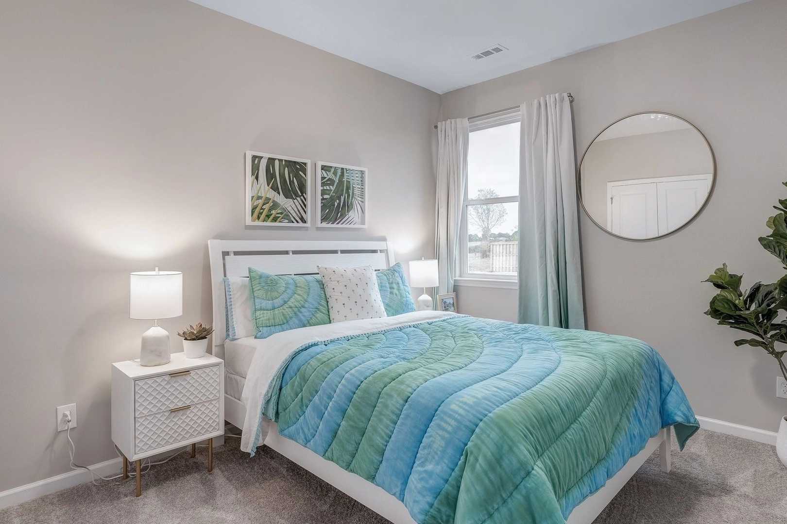 Spacious bedroom at Carroll Green in Harvest AL by Evermore Homes with queen bed teal-green bedding white nightstands round mirror and potted plant
