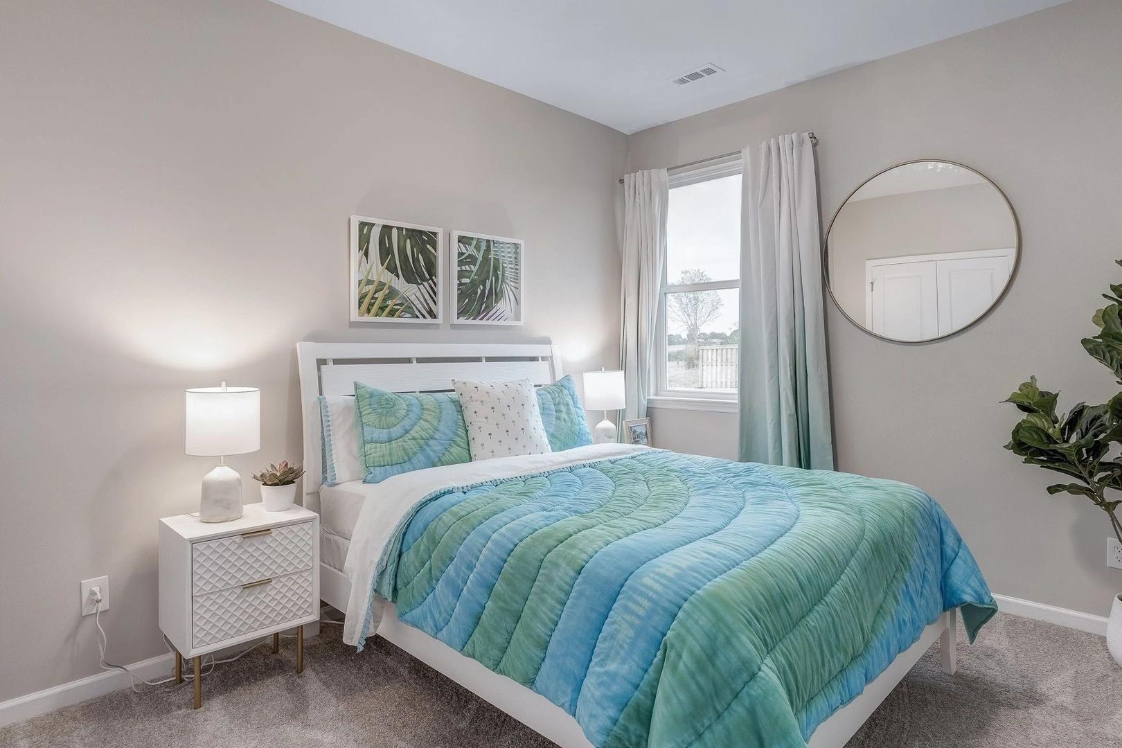 Spacious bedroom at Carroll Green in Harvest AL by Evermore Homes with queen bed teal-green bedding white nightstands round mirror and potted plant