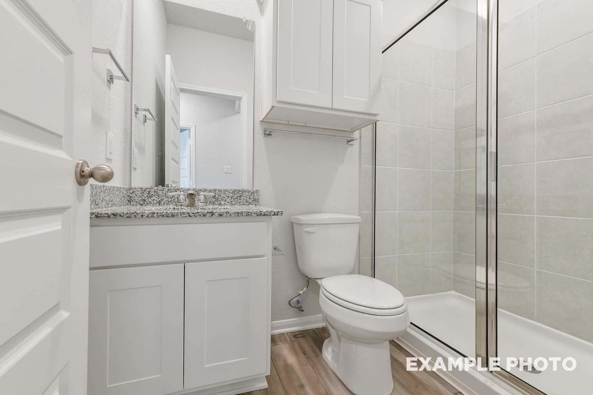 Modern bathroom in The Frio floor plan featuring a granite vanity, white cabinetry, and a glass-enclosed walk-in shower.