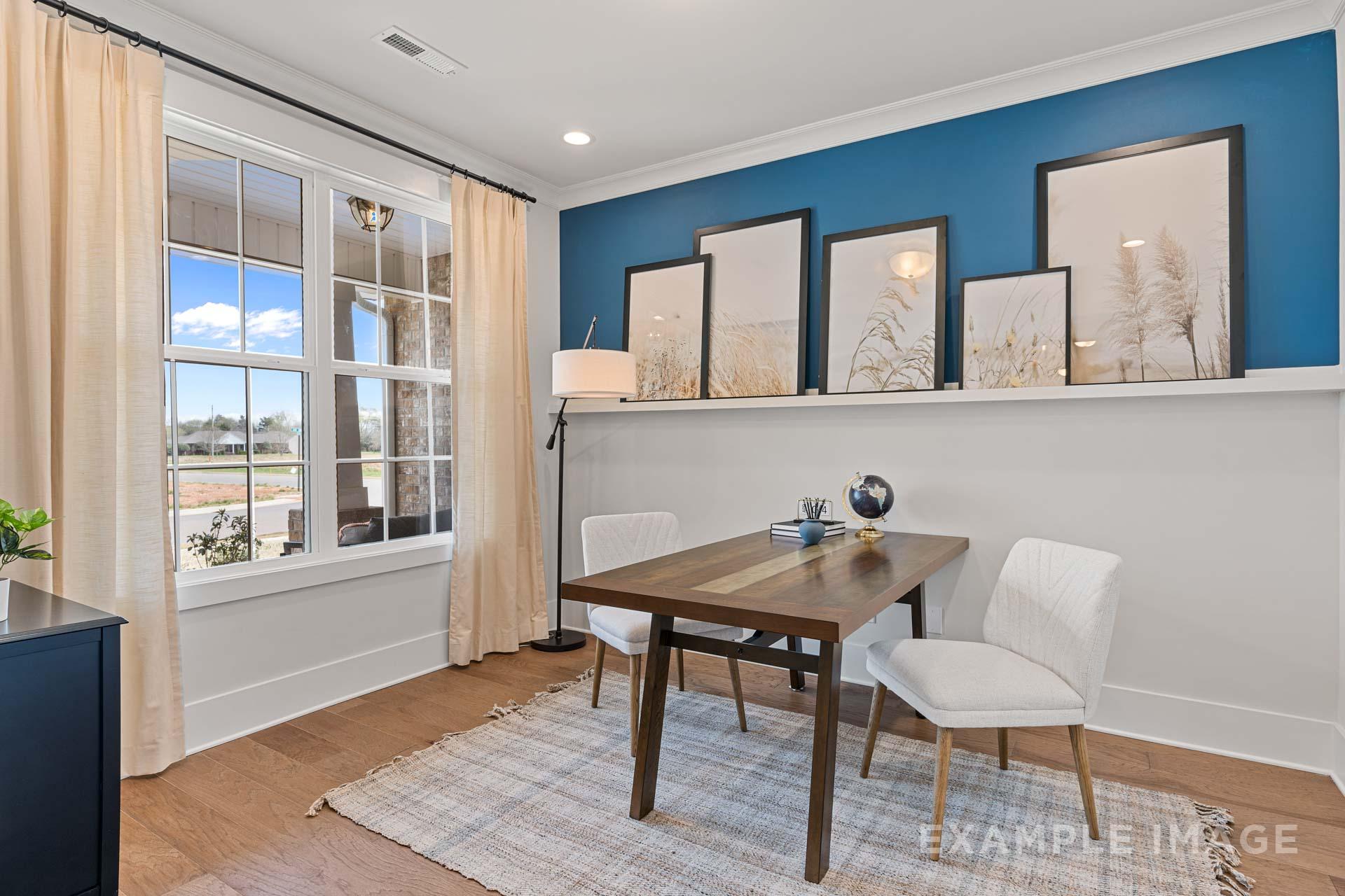 Spacious home office in The Rockford with blue accent wall, wooden desk, white chairs, and large windows overlooking fields