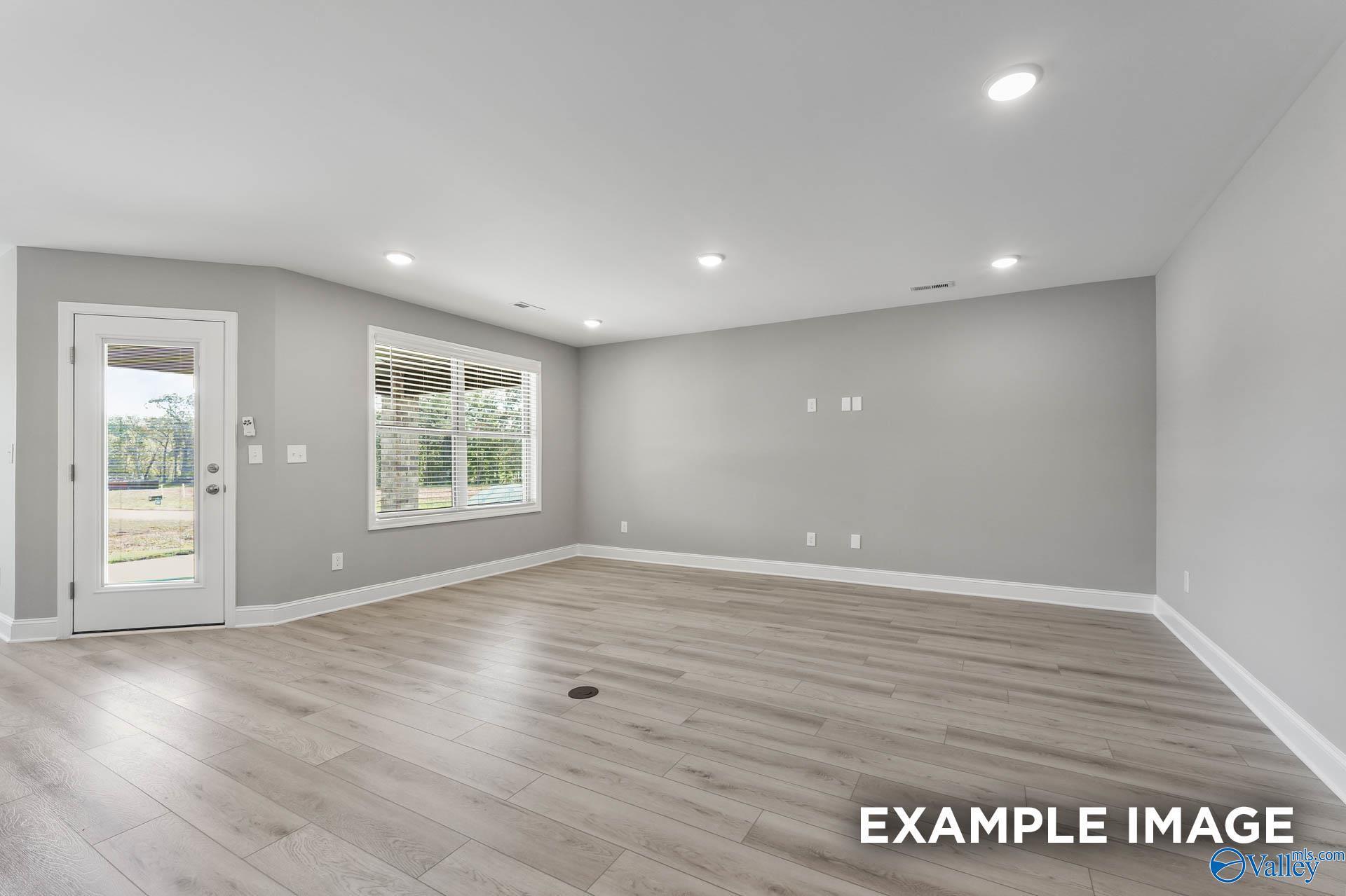Empty family room with gray walls, light wood laminate floors, large window views of trees, and glass door to patio in Davidson Homes Everett B, Toney AL
