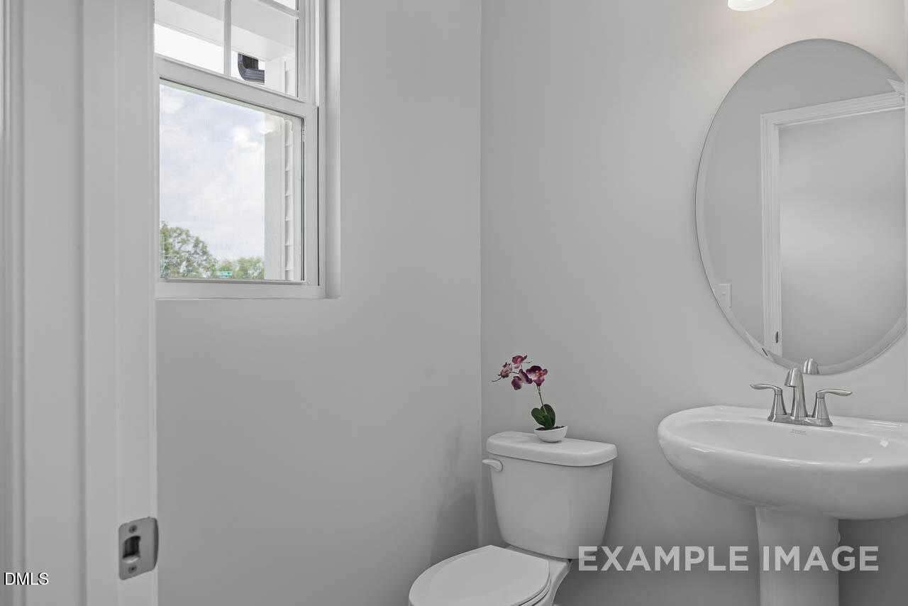 Bright powder room with white sink, round mirror, toilet, and window overlooking trees in Davidson Homes The Warren, Fuquay-Varina