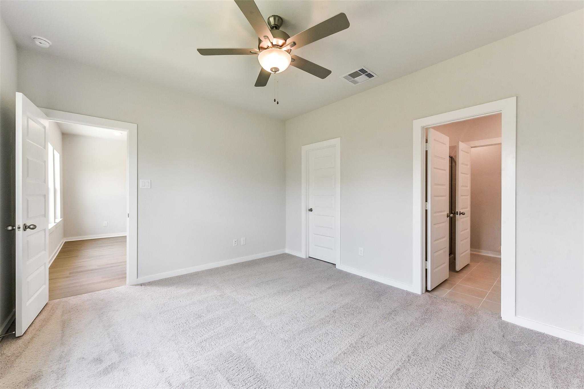 Bright bedroom with ceiling fan, beige walls, carpet floor, and doors to bathroom and closet in Davidson Homes The Frio F, Dayton Texas